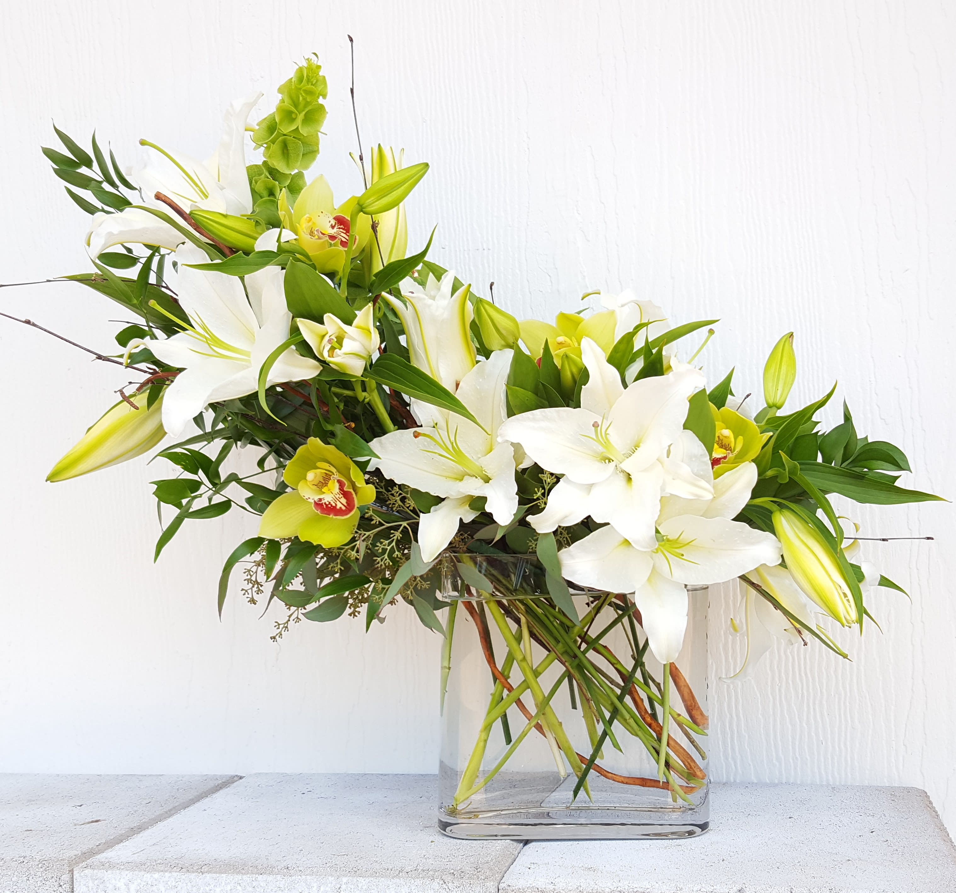 Contemporary lilies and orchids - Beautiful cascade of cymbidium orchids and white Casablanca lilies. Measures approx.  28" wide x 17" tall. As shown this arrangement is one sided,  deluxe makes it all round and premium is all round and in a taller, bigger vase. Florals and Glass container can vary depending on availability.