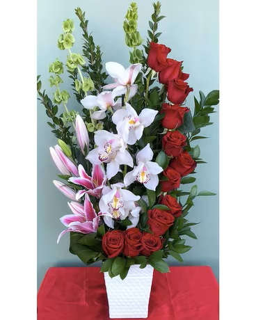 Majestic Symphony  - The "Majestic Symphony" is a deluxe floral arrangement that captures the essence of elegance and sophistication. This stunning display features a cascade of vibrant red roses, symbolizing love and passion, gracefully flowing through the arrangement. Interwoven among the roses are exquisite oriental pink lilies, adding a touch of exotic beauty and a delightful fragrance.  The Bells of Ireland contribute a unique texture and a refreshing pop of green, creating a striking contrast against the rich hues of the flowers. Cymbidium orchids, known for their elegance and charm, are thoughtfully placed to add a layer of sophistication and allure.  All these beautiful blooms are artfully arranged with fresh greens, enhancing the natural beauty and providing a lush backdrop. The arrangement is presented in a sleek glass container, allowing the viewer to appreciate the full depth and dimension of the design.  "Majestic Symphony" is perfect for making a grand statement at any event or as a luxurious gift to express heartfelt emotions. Its harmonious blend of colors and textures creates a visual masterpiece that is sure to captivate and inspire.