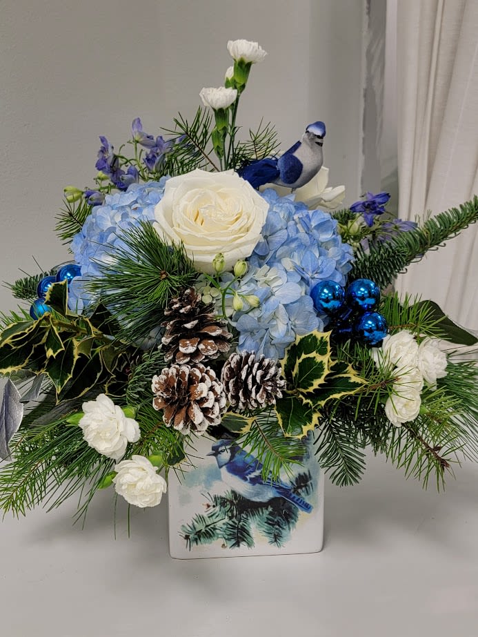 Winter Blue Jay - A beautiful white ceramic cube decorated with a lovely Blue Jay in a winter setting. Flowers include Blue hydrangea, white roses, white mini carnations, mixed winter greens, pinecones and blue Christmas balls. Includes a mini size Blue Jay. Standard is shown, Deluxe and premium sizes include additional stems and the arrangement increases in size.  Container is 5x5, Standard arrangement is 12x14.