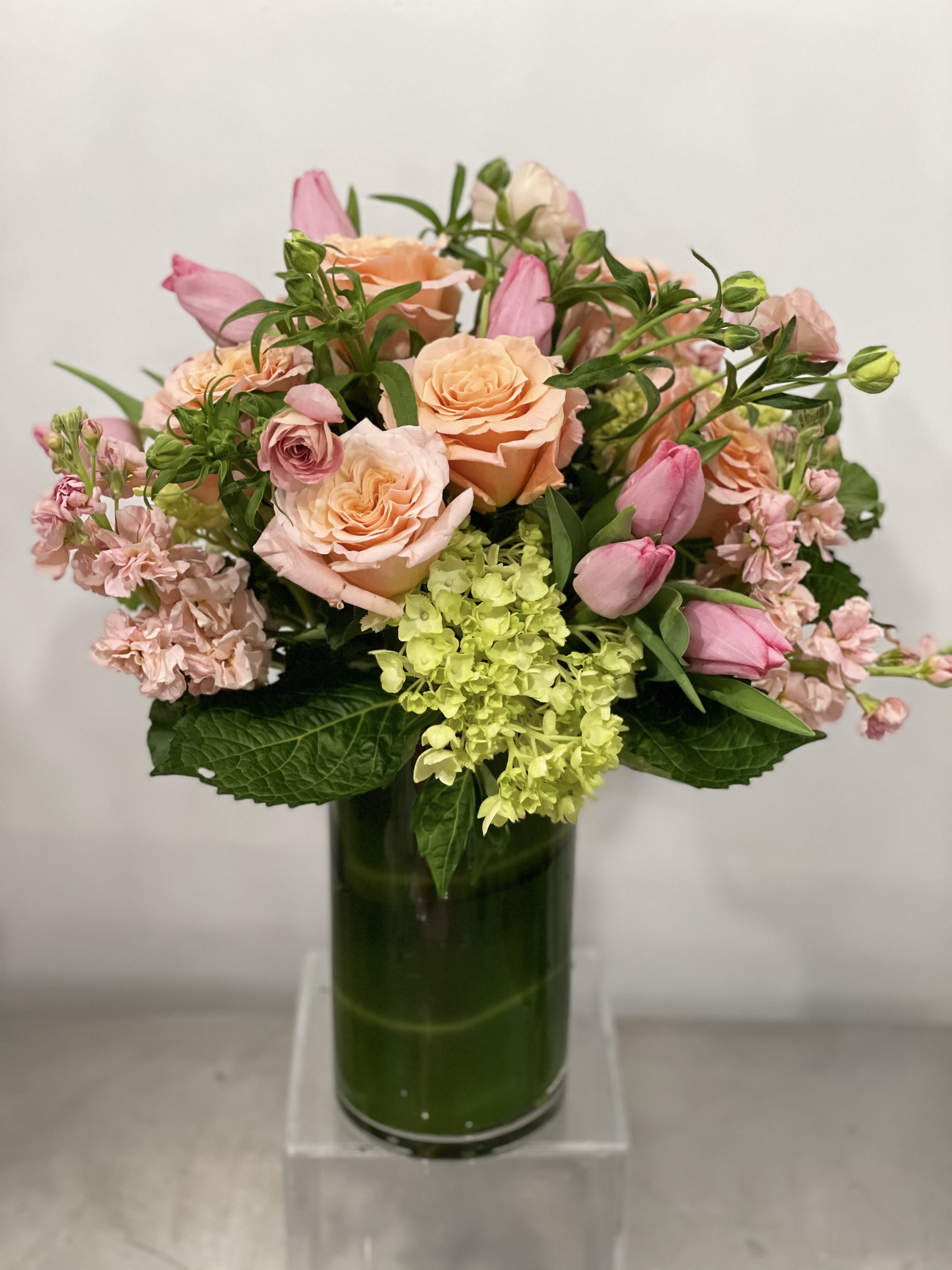 Pretty and Peach - Tall arrangement of pink and peach florals make for a lovely arrangement for a kitchen counter, foyer table, or credenza. Sweet smelling stock, fluffy hydrangea, ruffly roses, butterfly ranunculus, and spray roses in a tall 10" ti leaf lined vase. The picture is meant to represent they style and color palette of what we will send out on your behalf.  NOTES: We will choose the freshest and most interesting ingredients at the market to make this arrangement shine. Vessel may be substituted depending on availability.   If any design element such as color or flower/foliage are very important to your order please contact us to ensure availability first by calling the shop at  415 751 2747 