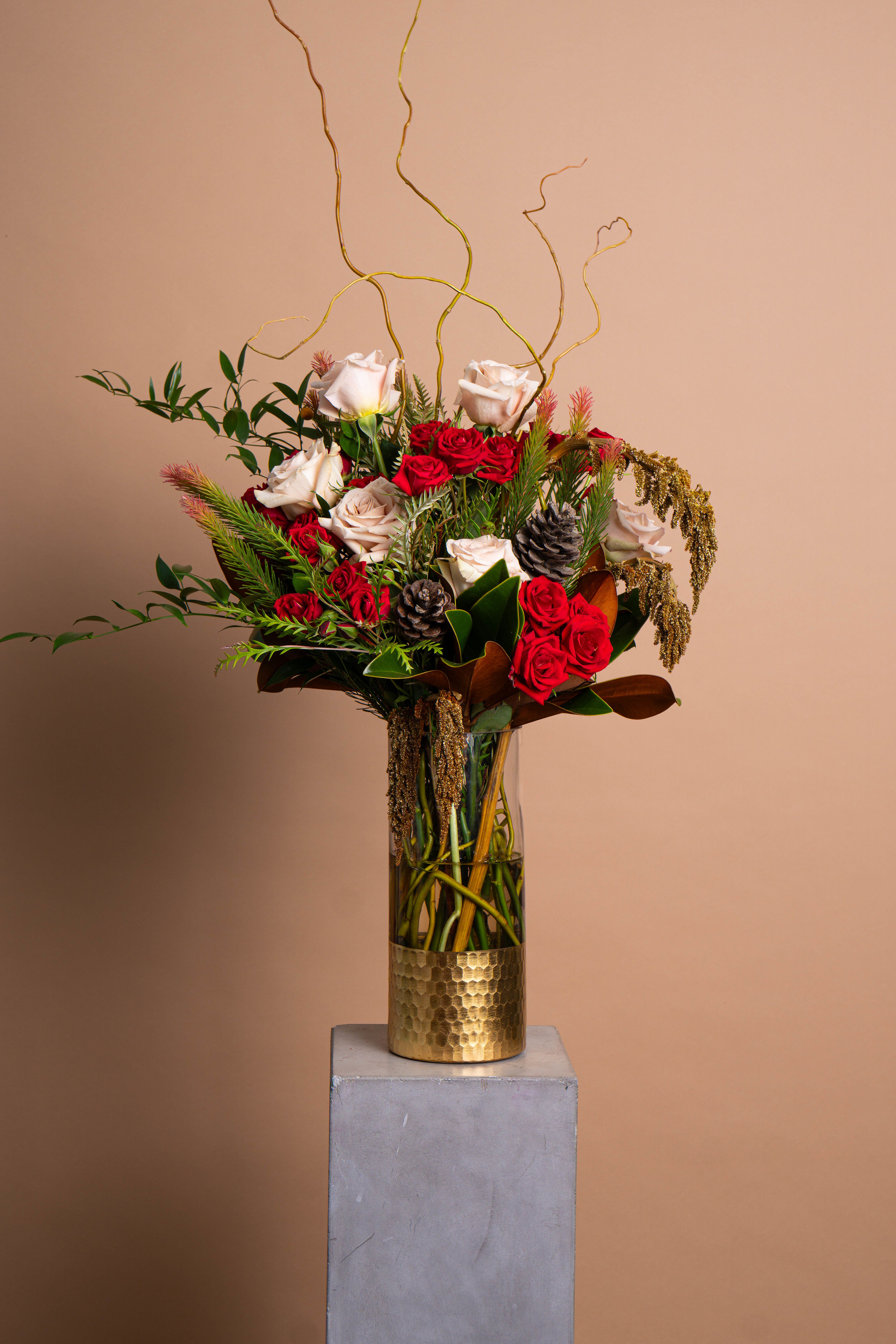 SCARLET &amp; PINE - A modern take on seasonal beauty. SCARLET &amp; PINE blends blush and crimson roses with textural evergreens, golden amaranth, and sculptural pinecones. Curving branches rise above the arrangement, creating a sense of movement and elegant height.  Warm, refined, and quietly festive, SCARLET &amp; PINE captures the spirit of the season through texture, light, and tone.  The arrangement pictured is Deluxe.