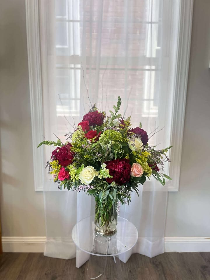 Love's Garden - Overflowing with timeless romance and natural beauty, Love’s Garden is a lush, garden-style arrangement that speaks straight to the heart. This enchanting design features velvety burgundy peonies, classic red roses, soft blush and ivory roses, and fresh green hydrangea, all nestled among sprigs of solidago, waxflower, and cascading eucalyptus.  Arranged in a tall clear glass vase, this piece combines rich color with graceful texture, making it perfect for expressing deep love, heartfelt sympathy, or marking a truly special occasion. The blend of soft pastels and dramatic florals creates a balanced and breathtaking tribute to the beauty of love in full bloom.