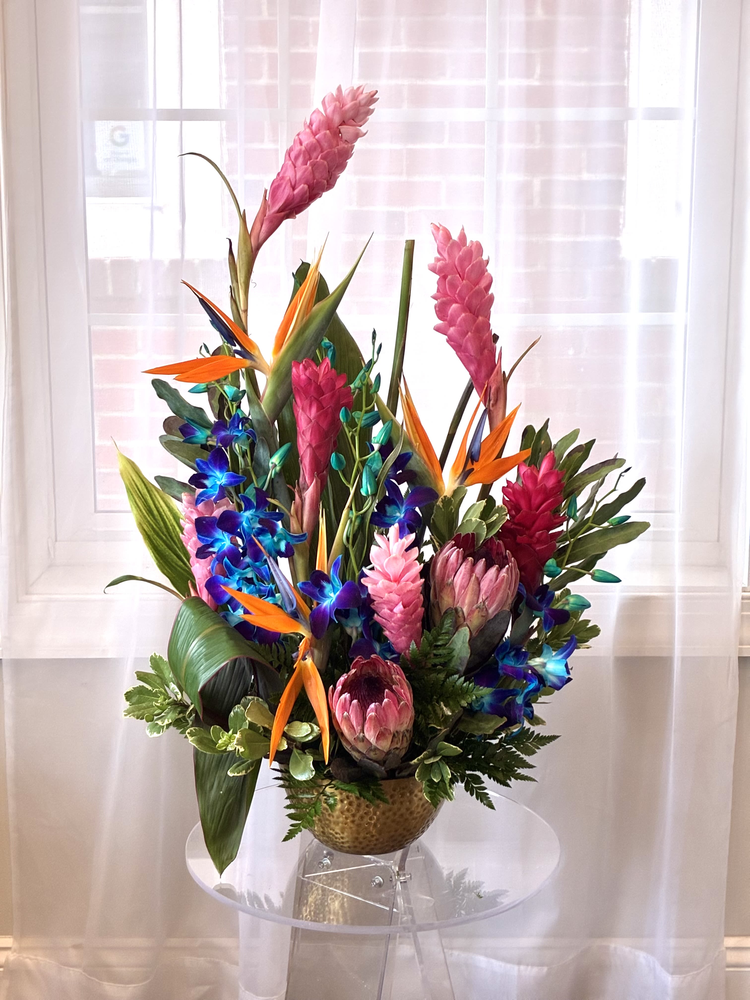 Tropical Treasure Luxe Arrangement - Transport someone straight to the islands with this eye-catching showpiece bursting with exotic blooms and bold color Towering pink torch gingers and velvety king protea deliver sculptural drama. Electric-blue dendrobium orchids and fiery birds of paradise add vibrant pops of teal, orange and indigo.  Lush tropical foliage—monsteras, palm blades and glossy greens—frames the flowers in rich texture.