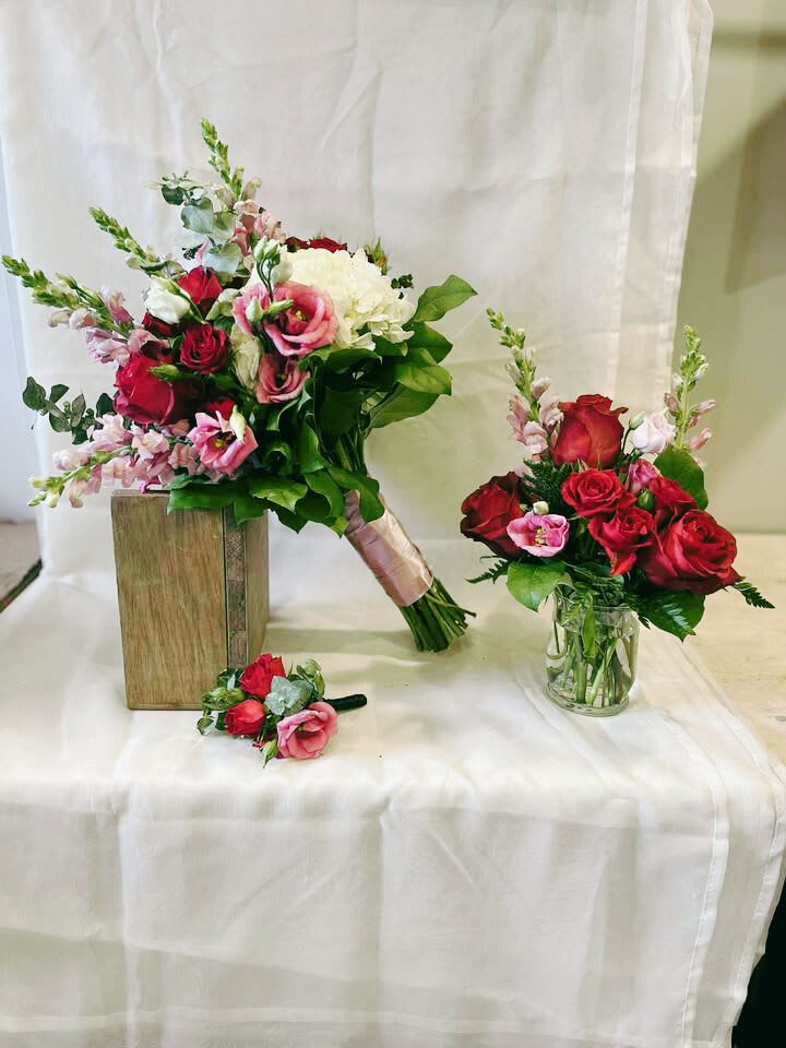 "Wine &amp; Whispers" Wedding Floral Collection - A captivating trio designed for the elegant and romantic bride. Featuring rich red roses, delicate mauve lisianthus, soft snapdragons, and creamy hydrangea, this set includes:  Bridal Bouquet: A stunning hand-tied bouquet overflowing with lush florals, wrapped in blush satin ribbon for a soft, elegant finish.  Boutonnière: A romantic touch of coordinated blooms and greenery for the groom or groomsmen.  Mason Jar Arrangement: A charming, petite arrangement ideal for cocktail tables or side décor.  Feature Centerpiece: A grand, full-bodied arrangement in a clear cube vase—perfect for the couple’s table, guest tables, or as a ceremony focal point. It features a bold blend of red roses, creamy hydrangeas, and textured greenery, creating an unforgettable statement.  Perfect for wine-country weddings, vintage-inspired ceremonies, or any celebration filled with romance and timeless beauty.  *Please check individual pricing structures for what is included*