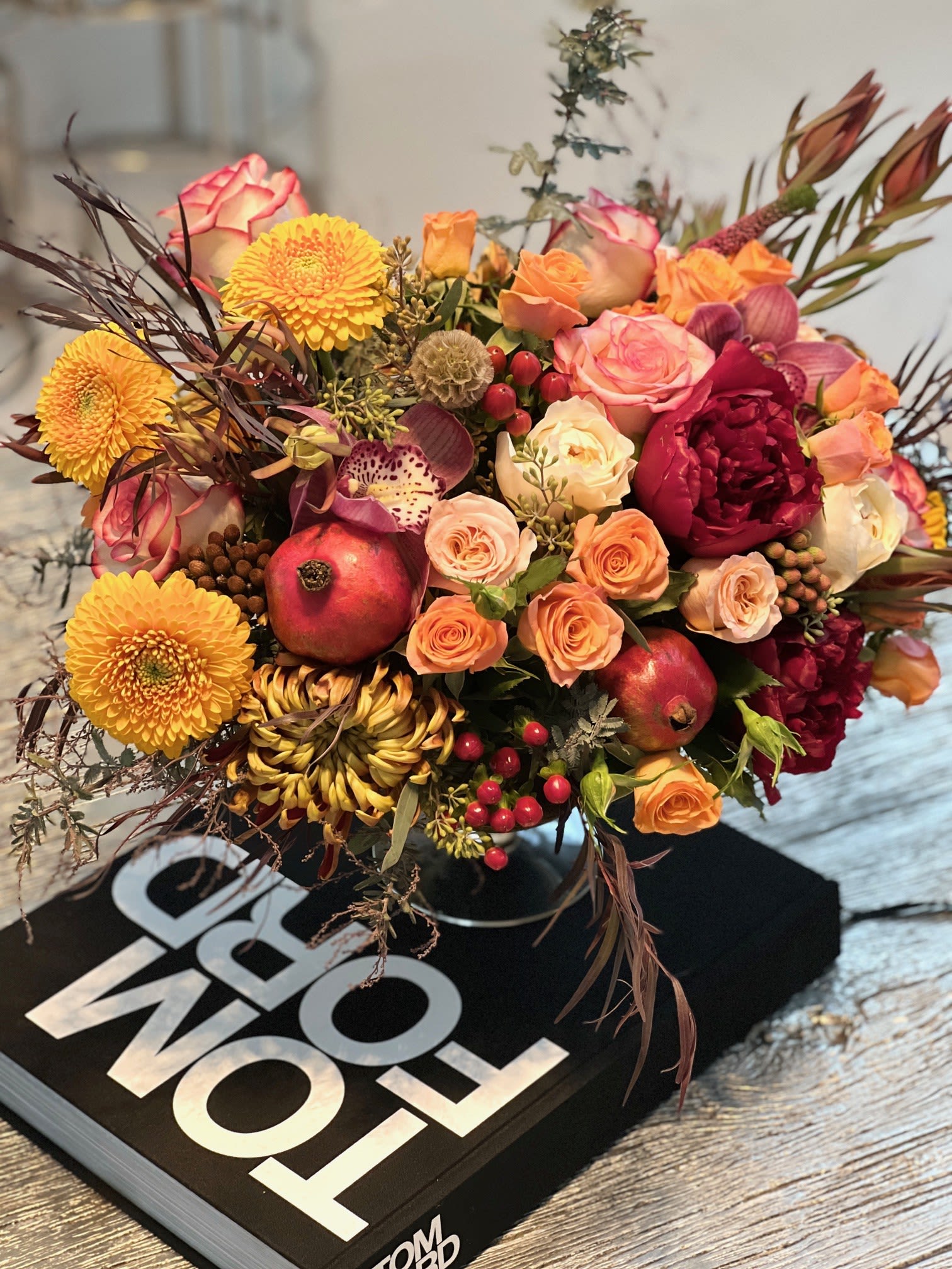 Autumn Garden ™ - This opulent centerpiece bursts with autumnal charm, blending rich burgundy roses, golden chrysanthemums, and peach spray roses with textural accents of berries and wispy branches. Nestled among the blooms are pomegranates for a lush, seasonal touch, creating a warm and abundant display. Perfect for a sophisticated tablescape or gift, it radiates a cozy harvest feel with a luxurious edge.