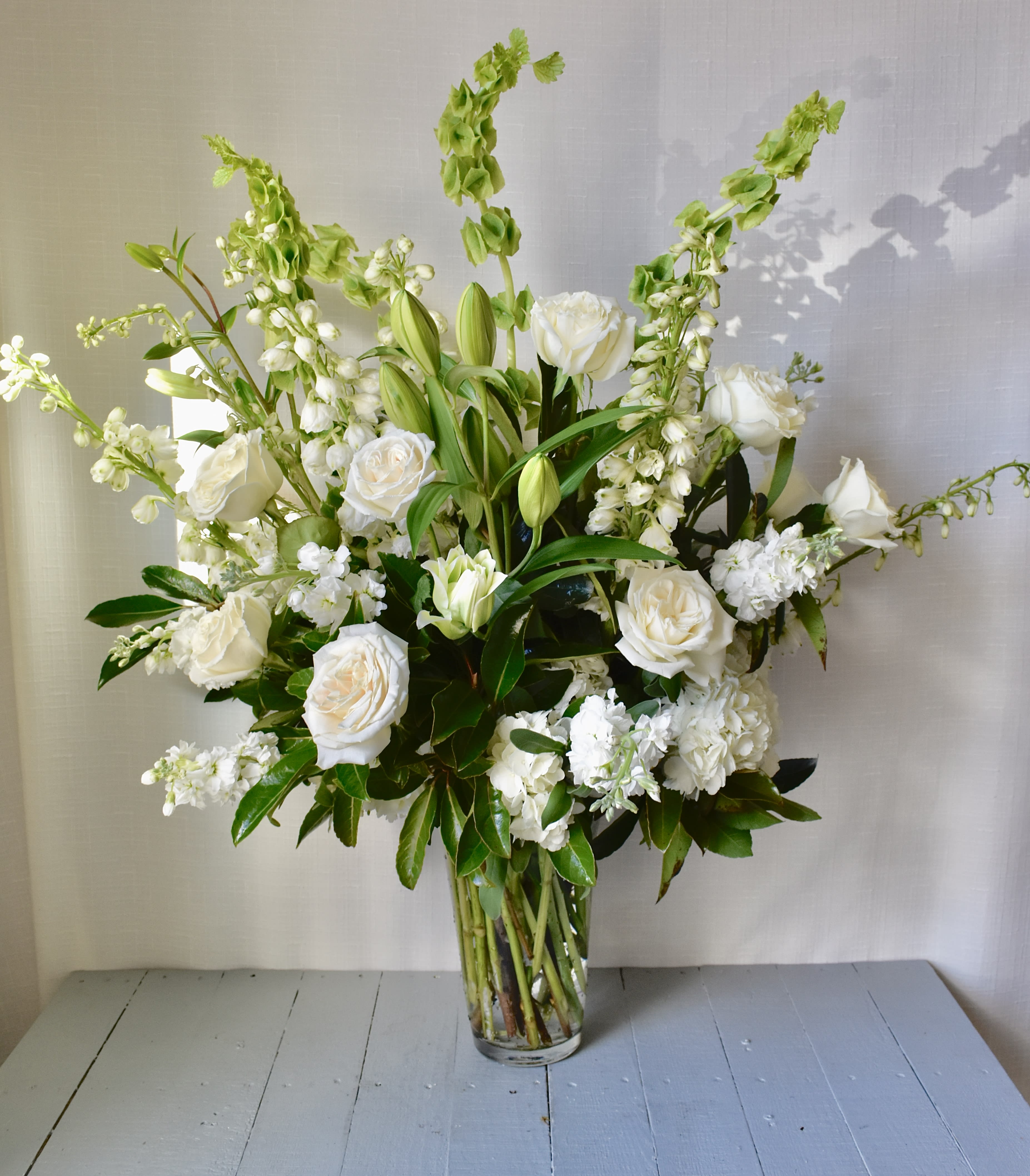Heartfelt Wishes - Make a clean, impactful statement with this elegant, all white floral design. "Graceful Wishes" features all white roses, bells of Ireland, and stock on a bed of vibrant foliage.  Flowers may vary depending on availability and season.