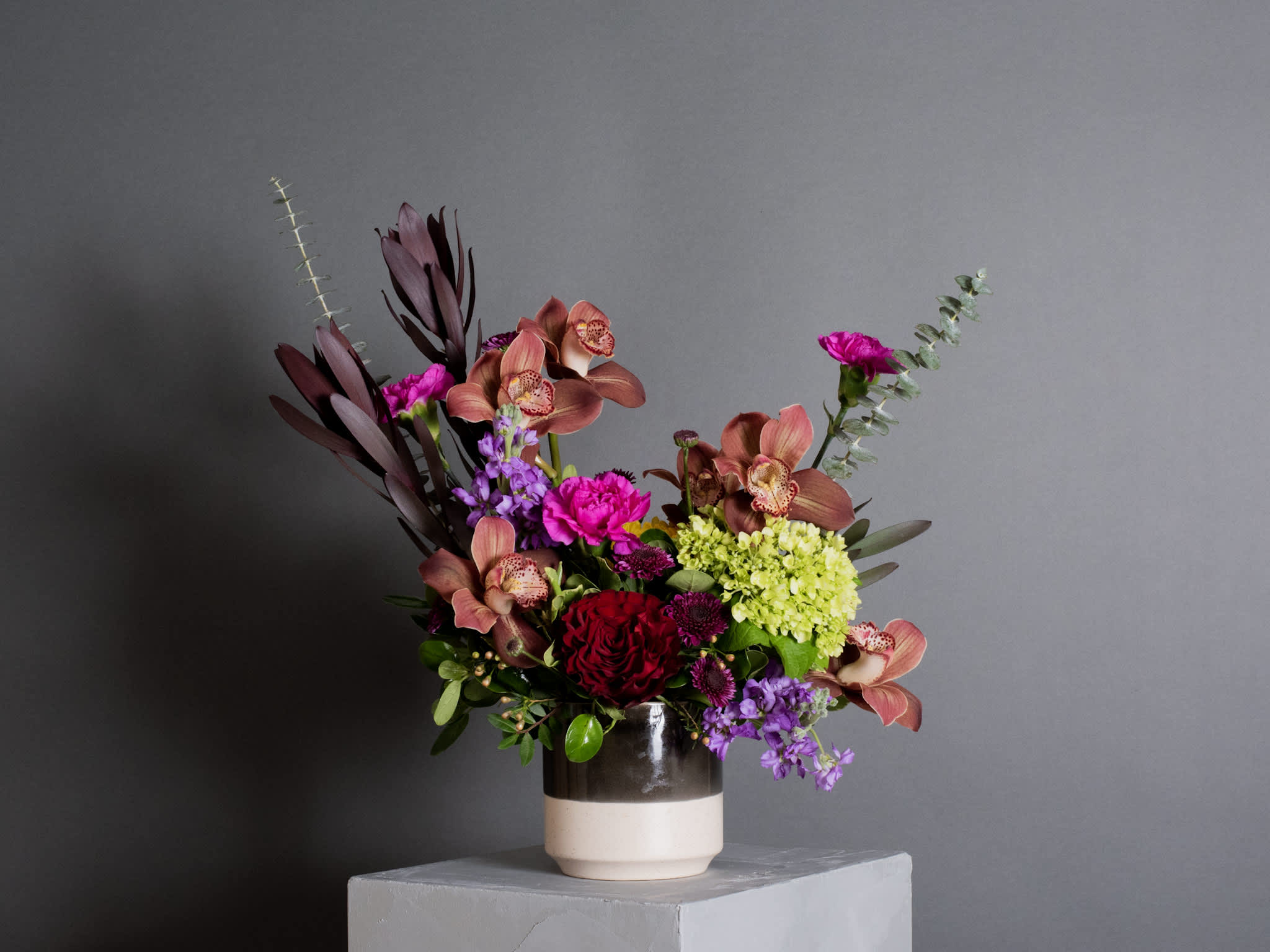 London, Wanderlust Series - Ceramic Pot Arrangement - A striking arrangement inspired by London’s vibrant energy, featuring dramatic colors. This design showcases elegant cymbidium orchids, lush eucalyptus, soft mini hydrangea, a romantic heart rose, and bright pink carnations, creating a truly captivating display.  Product shown at $134.95  Pot size : 4.5in X 4.5in