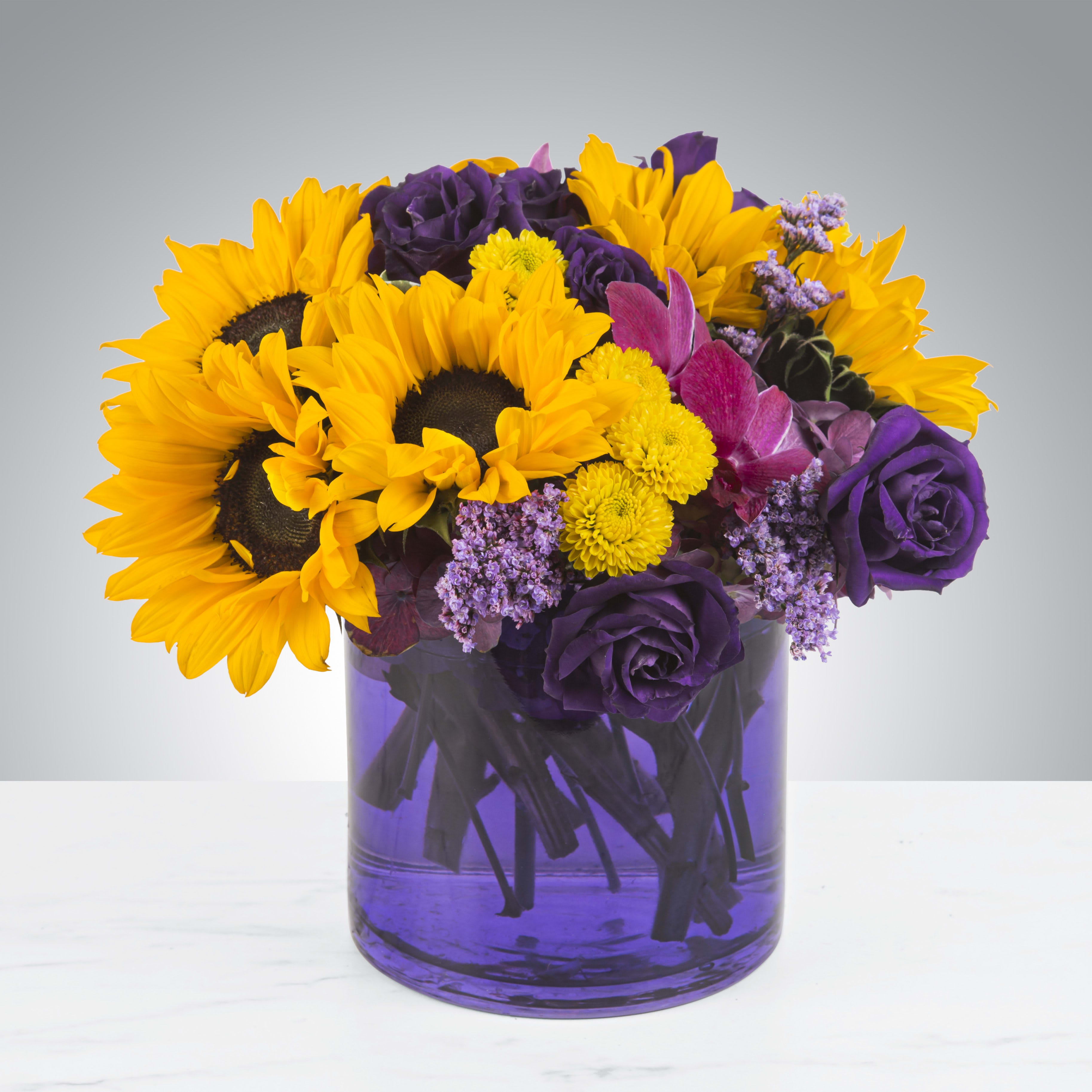 Make Some Magic by BloomNation™ - Celebrate Mardi Gras year round and throw some beads on it with this fun and colorful arrangement.  Purple lisianthus and roses come together with sunflowers and mini mums to create a bright piece in a purple vase.    APPROXIMATE DIMENSIONS 11" W X 11" H
