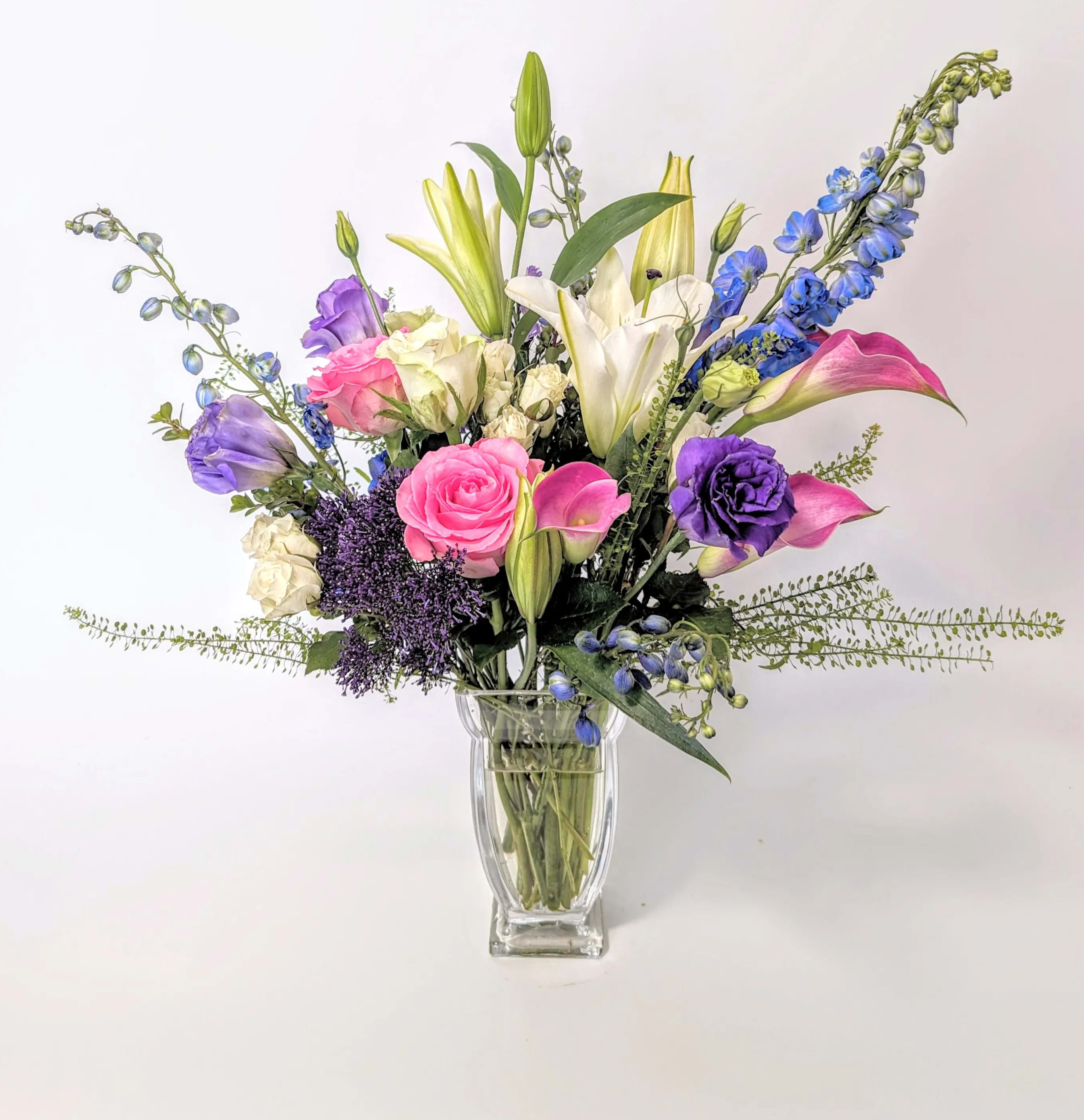 Soft Caress - A beautiful mixed bouquet of the freshest premium flowers available like, pink roses, blue delphinium, cala lilies, spray roses, monte casino, lisianthus, white lilies and more all delicately arranged  with wispy greens in a beautiful high class clear glass couture vase. A true statement of beauty and elegance and perfect for all occasions. Make this bouquet extra special and add on a fragrant candle or hand cream to complete your gift.   Measures approx. 20" high and 18" wide