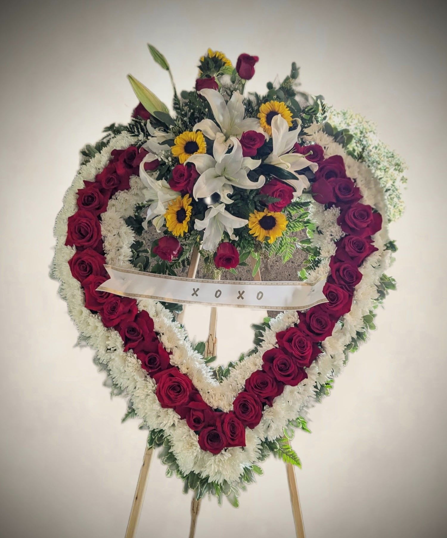 Heartfelt Sympathy Wreath  - A graceful heart-shaped sympathy wreath designed with deep red roses bordered by soft white blooms, symbolizing love, remembrance, and peace. The center arrangement features elegant white lilies accented with cheerful sunflowers and lush greenery, creating a meaningful contrast of warmth and serenity. Displayed on an easel and finished with a ribbon reading “XOXO,” this tribute offers a heartfelt expression of love, comfort, and lasting remembrance. Ideal for funeral services, memorials, or celebrations of life. 