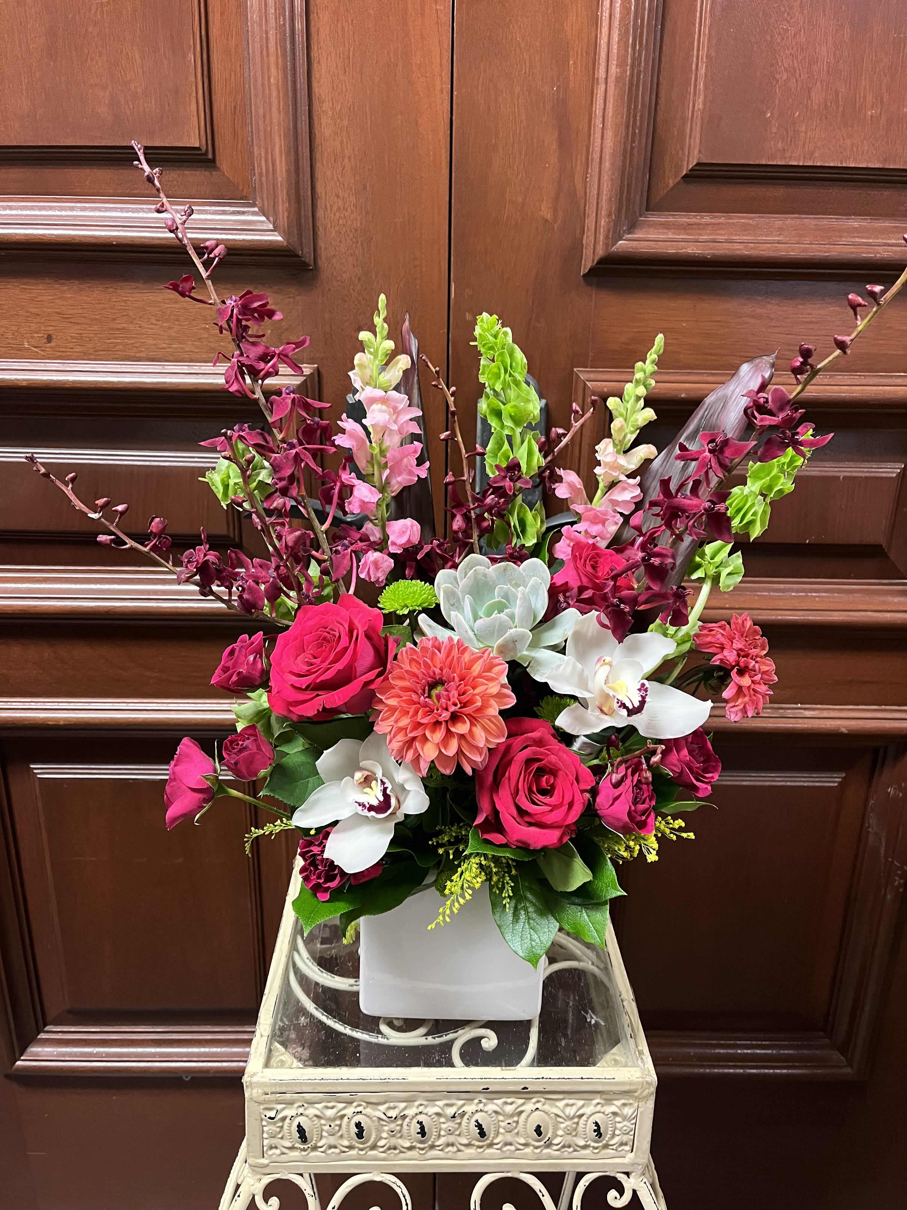 From My Heart to Yours - Recipe: Season Dahlias, Burgendy orchids, Cymbidium Orchid, Pink Snapdragon, Hot pink Roses, Red Spray Roses, Bells of Ireland, Succulent and more in a square, white vase. Design View: Symmetric Front Facing View Photo shown: Standard
