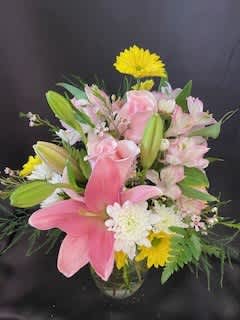 A Mother's Love - In a clear glass vase, Pink Lilies are surrounded by alstromeria, chrysanthamums, roses and wax flowers with seasonal greenery.