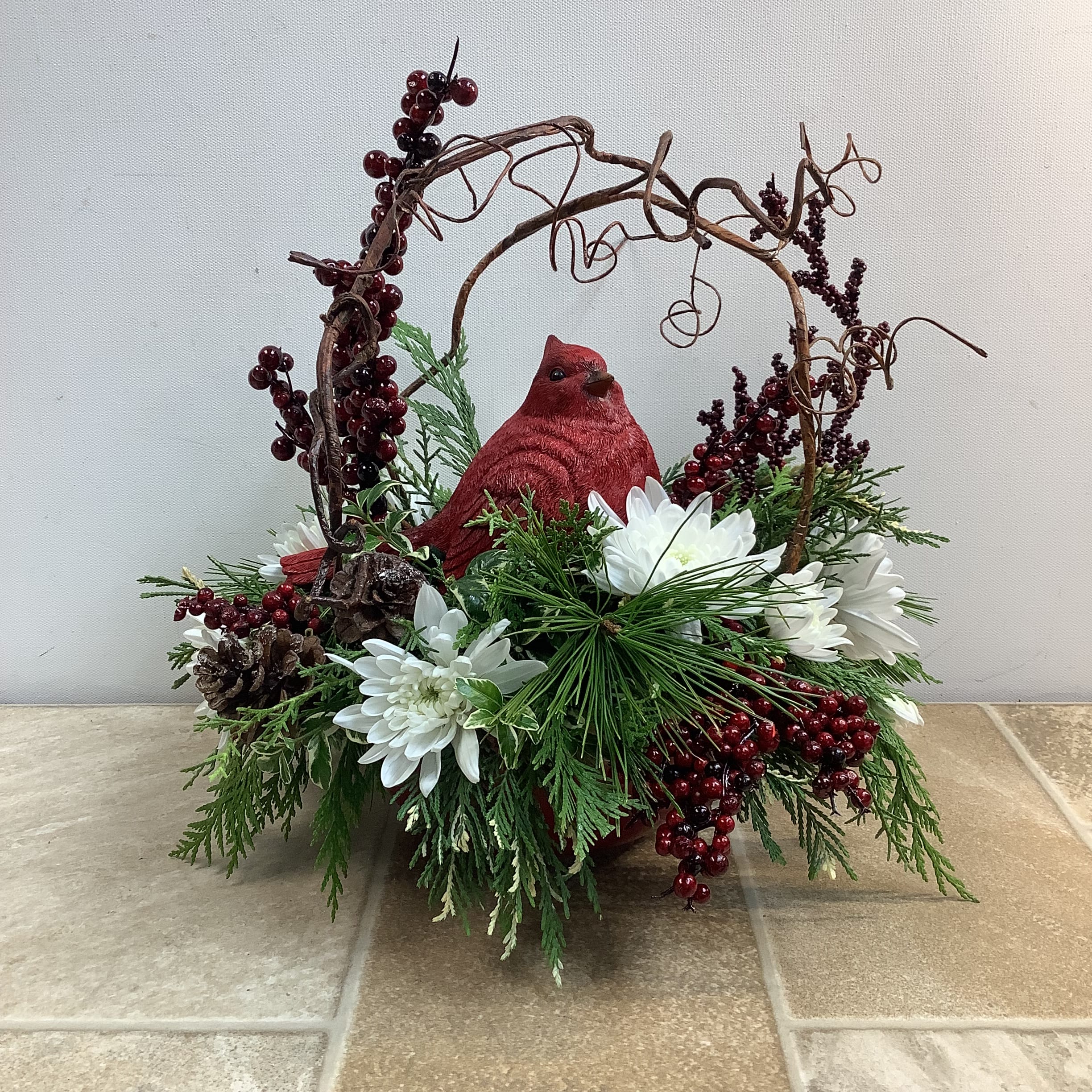 Rustic Charm - This long-lasting centerpiece is sure to make your holidays extra-special. Fresh evergreens, white chrysanthemums, silk berries pine cones, and a twisty brown artificial vine are beautifully arranged around a keepsake ceramic cardinal.   Primary flowers: mum, evergreen  Flower delivery La Crosse, Onalaska, Holmen, West Salem, La Crescent. Flower shop. Plant store. Best florist La Crosse Wisconsin area.  Seasonal and holiday floral creations. Funeral arrangements. New baby bouquets. Birthday celebration flowers. Get well arrangements. Just because gifts.