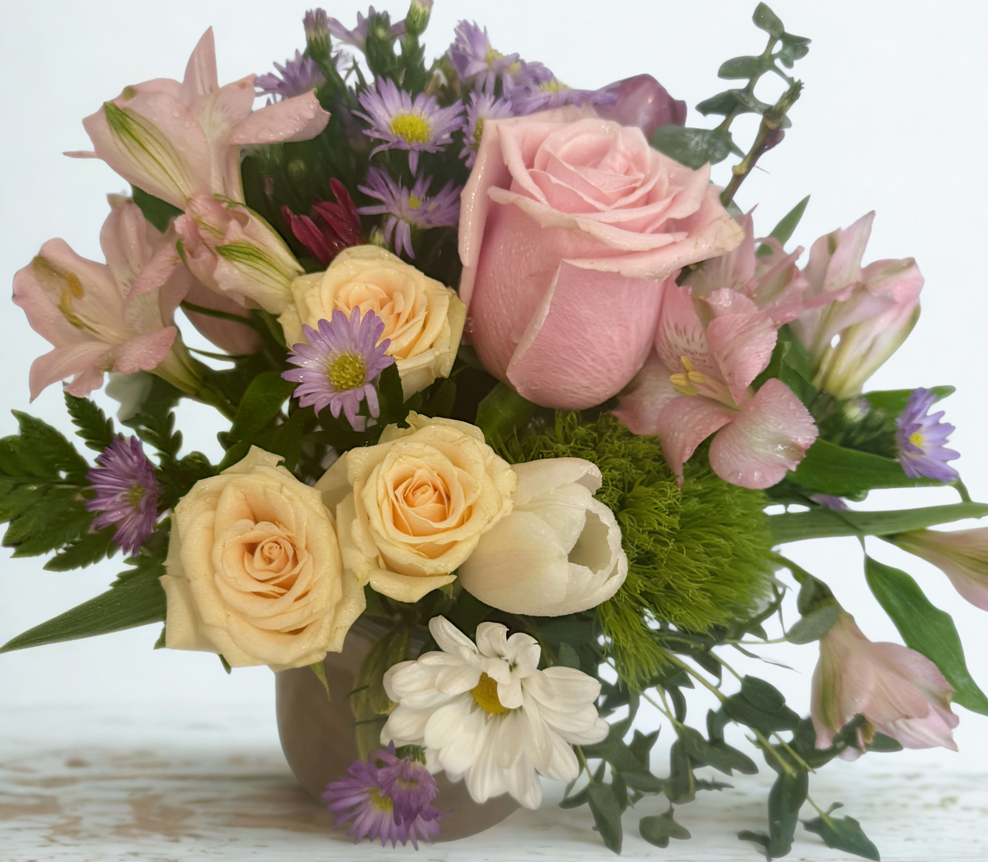 Pastel Charm - This delightful compact arrangement showcases a harmonious blend of soft pink and creamy yellow hues, accented by playful pops of purple and white. Nestled in a simple vase, it features classic roses, delicate alstroemeria, and charming aster-like blooms, creating a sweet and inviting display.