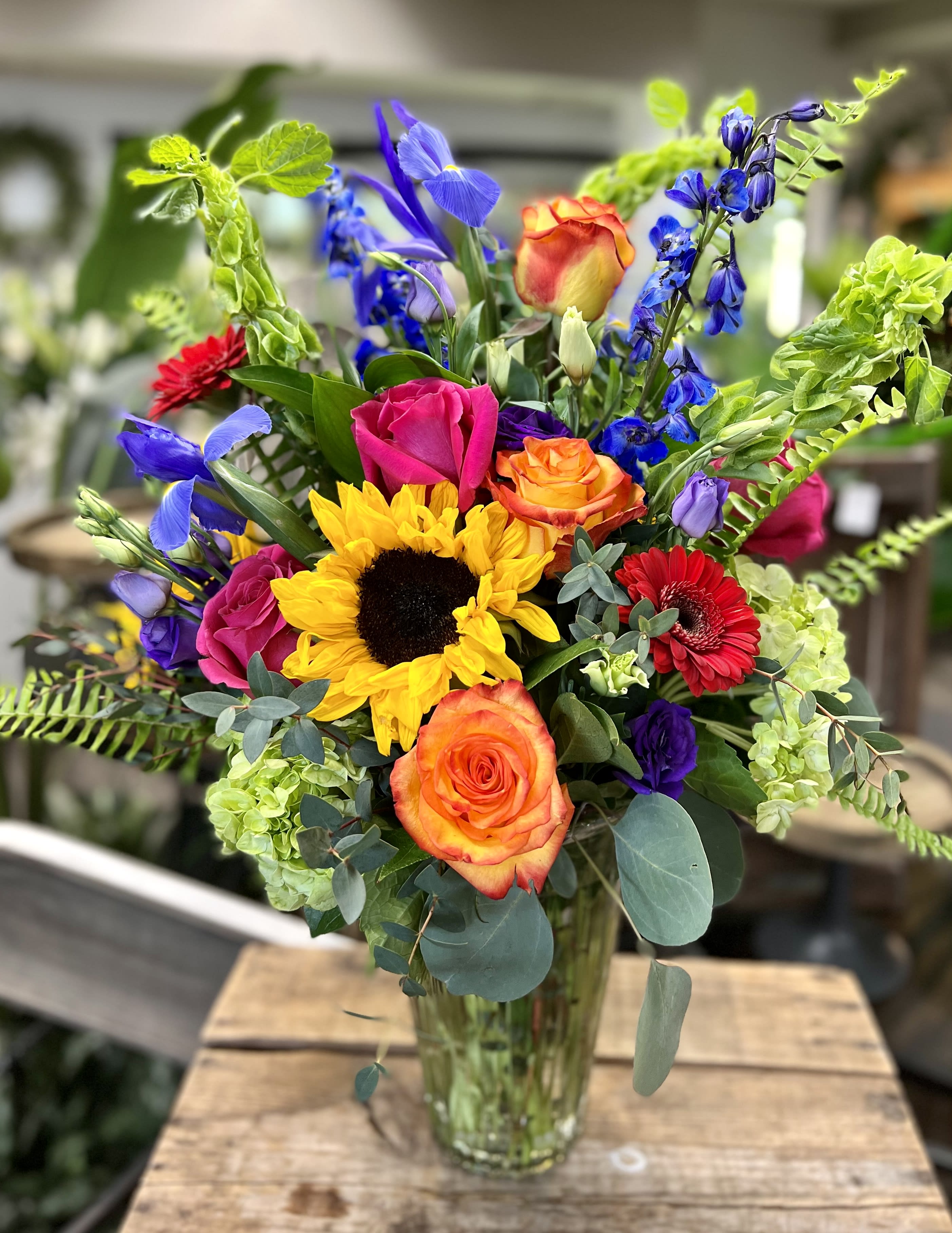 Vivid Jewel Bouquet - LOCAL DELIVERY ONLY!! A bright jewel tone fresh floral arrangement that includes; iris, sunflowers, gerbera daisies, hydrangea, delphinium, roses, bells of Ireland, Lisianthus and mixed greenery throughout. Designed in a clear glass vase. Flowers &amp; Colors may vary! AVAILABLE FOR LOCAL DELIVERY ONLY!!!