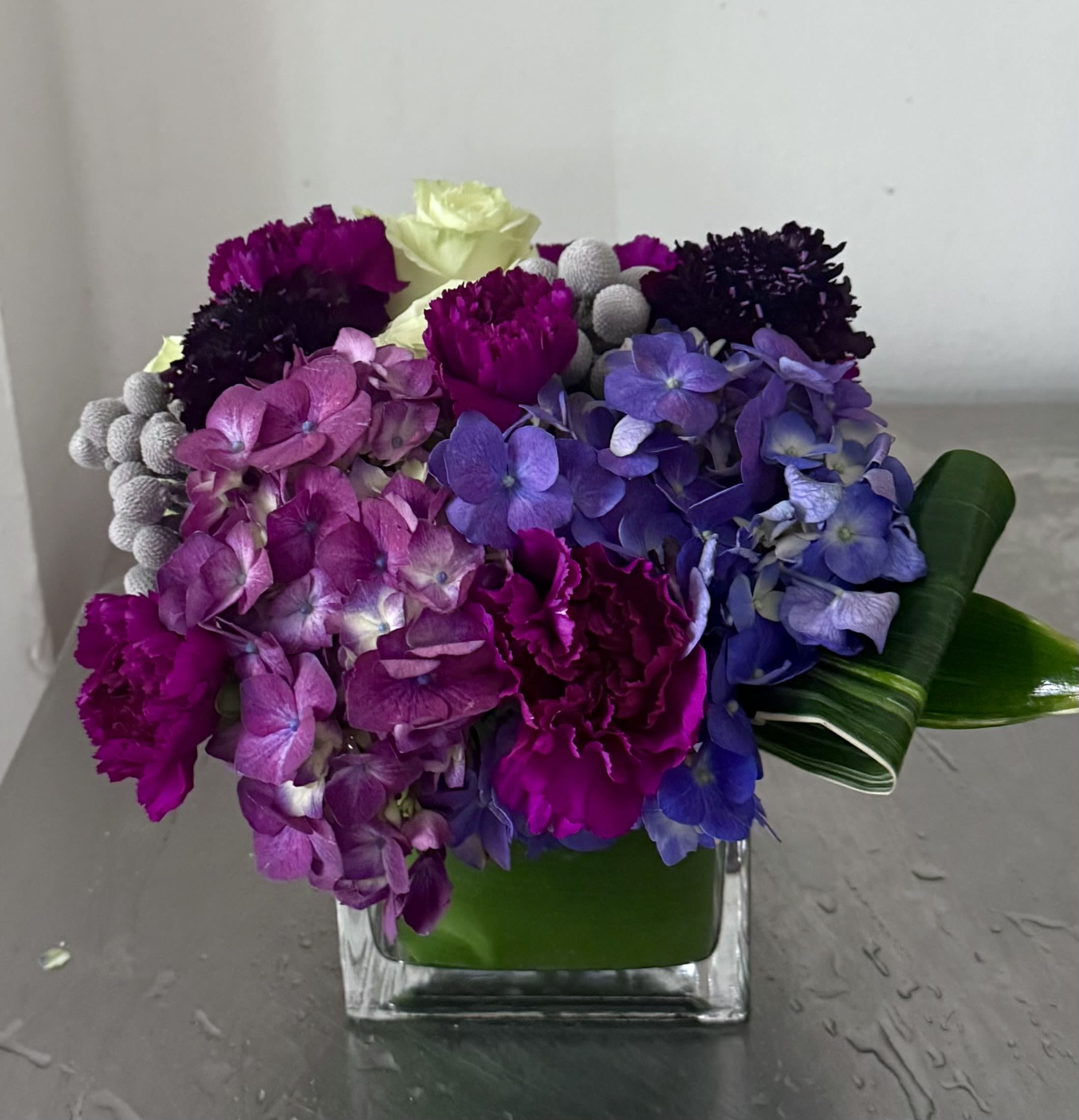 Ametyst Dream - The arrangement features a lush mix of deep purple carnations, dark pink hydrangeas, scabiosa and a touch of light green roses in 5” cube glass vase wrapped with a tie leaf.  