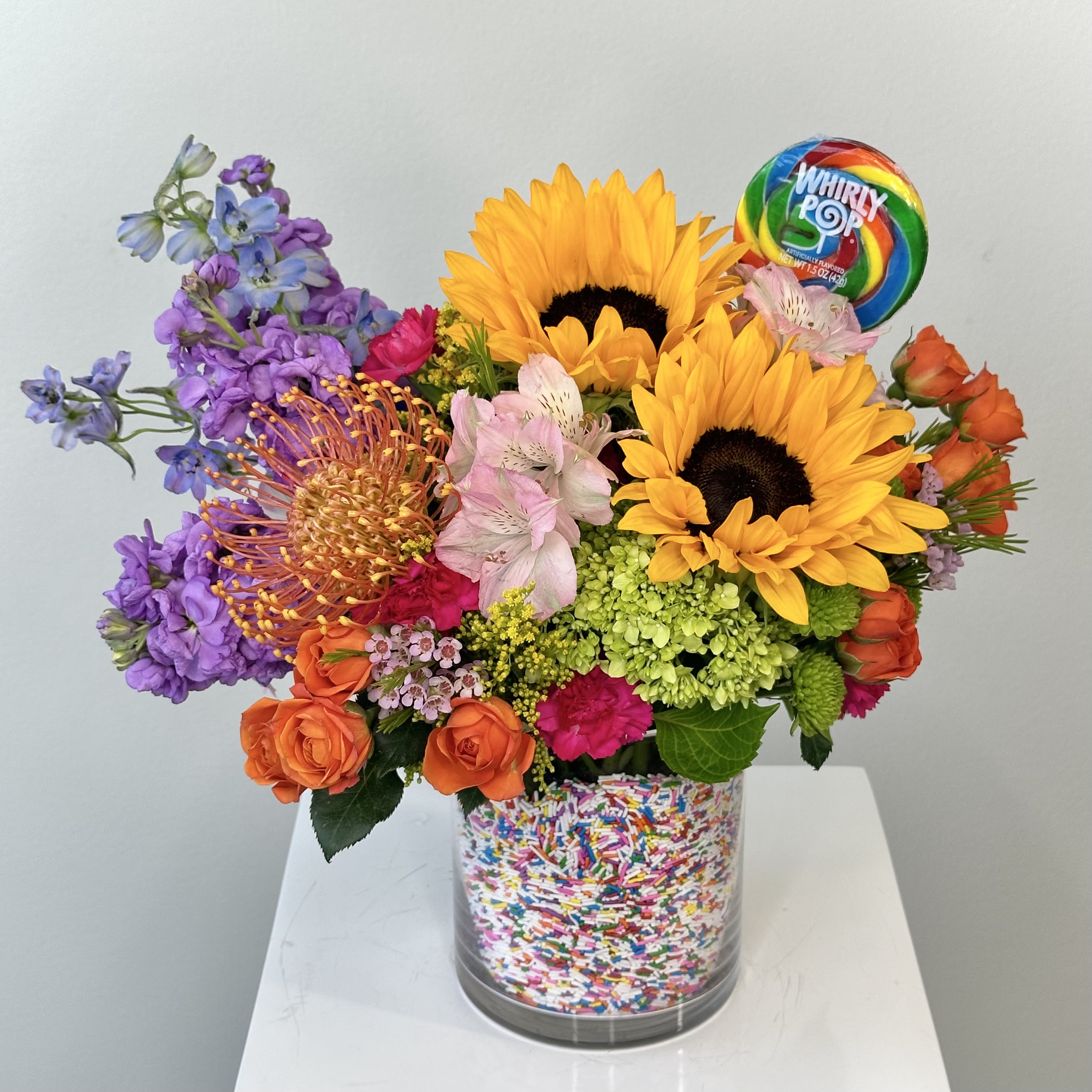 Sprinkles - Sprinkles is the ultimate burst of joy in a bouquet! A celebration of color and texture, this vibrant arrangement features bold sunflowers that instantly brighten any room, complemented by the delicate elegance of hydrangea and the whimsical charm of a pincushion flower. The soft petals of alstroemeria, cheerful carnations, and sweet spray roses add playful layers of beauty, creating a lively mix that’s as fun as it is stunning.  What really sets Sprinkles apart, though, is its unique presentation. The vase is lined with rainbow sprinkles, giving the arrangement a delightful pop of color and a fun, sweet touch. And as the perfect finishing touch, a bright, rainbow-colored lollipop is included, making this bouquet not only a feast for the eyes but a treat for the soul. Whether you’re celebrating a special occasion or just brightening someone's day, Sprinkles is sure to spread smiles and sunshine wherever it goes.