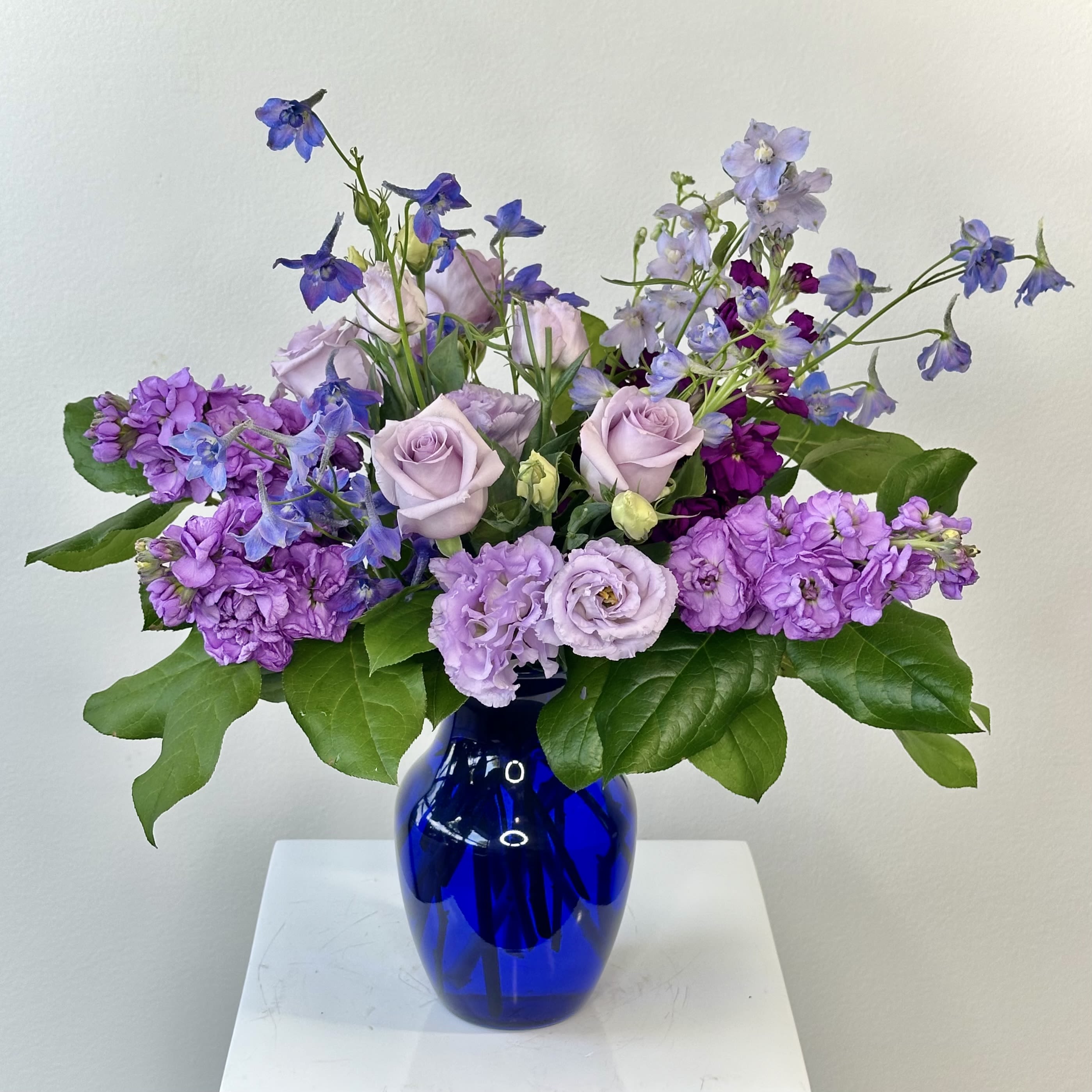 Twilight Glow Bouquet  - A whimsical treasure for dreamy hearts. Bathed in the soft blush of twilight, the Twilight Glow Bouquet from Irvine Village Flowers weaves a spell of enchantment with its delicate blue and purple blooms. This ethereal blend of velvety Delphinium, charming Stock, and tender Roses dances in hues of dusk, whispering sweet sentiments of congratulations, happy birthdays, or tender thoughts of you. Each petal glows with a rosy magic, perfect for wrapping your loved ones in a pink-tinted dream.    The Delphinium, with its starry spikes, shimmers like the evening sky, symbolizing open hearts and endless possibilities. The Stock, soft and fragrant, blooms with whispers of peace, affection, and timeless beauty. And the Roses, kissed by twilight’s glow, unfurl in a gentle embrace of love, joy, and admiration. Together, they craft a bouquet that’s as breathtaking as a pastel sunset, a gift to make any soul feel cherished.    The Deluxe Bouquet rises to a dreamy 17"H x 16"W, a vision of elegance and grace. Visit Irvine Village Flowers online or drift into our store to bask in this twilight fantasy. With our seamless flower delivery across Irvine, your magical creation arrives fresh, blooming with love and ready to sprinkle joy like stardust.