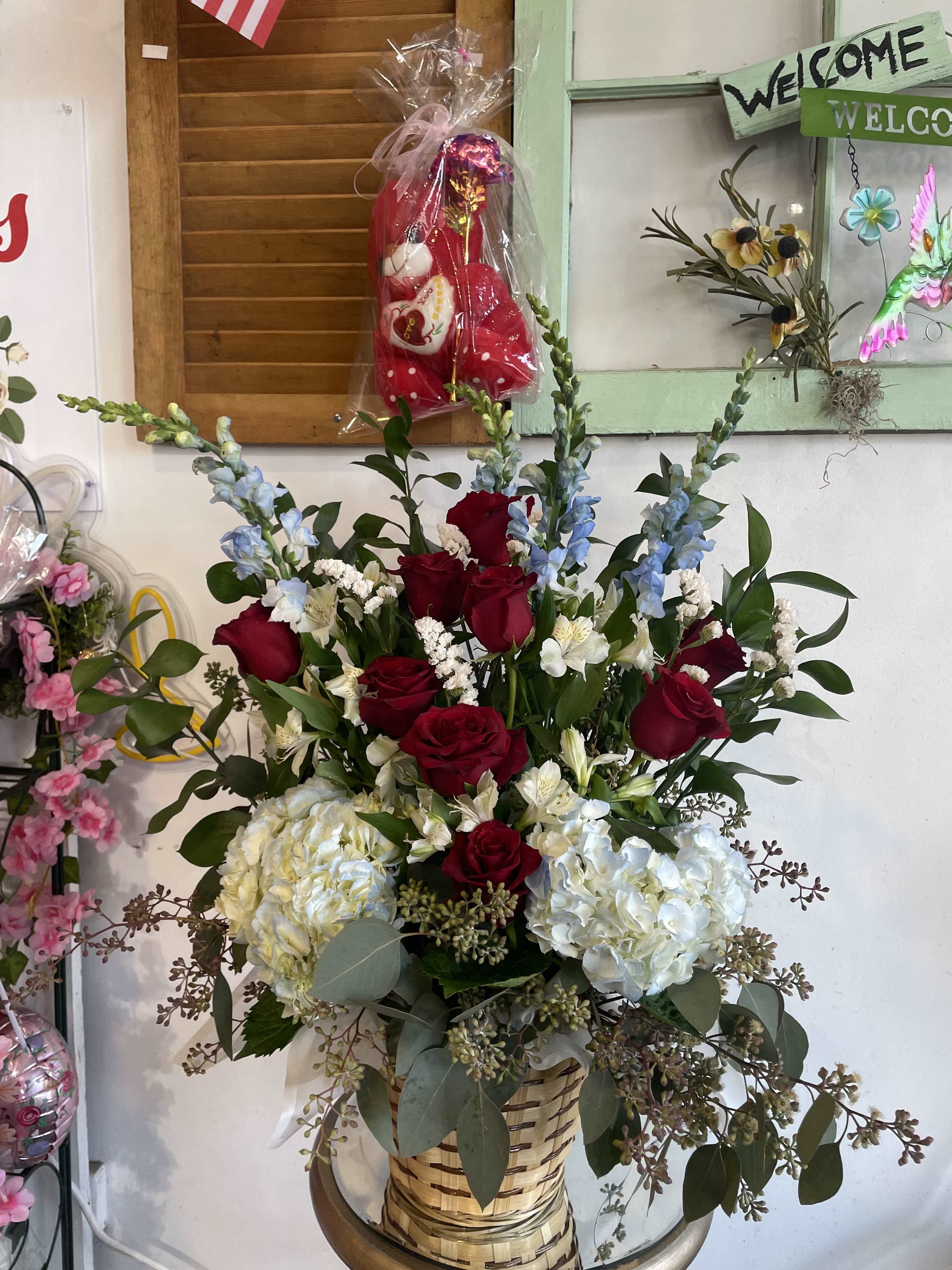 Garden of Whispered Love - A graceful blend of deep red roses, snowy hydrangeas, soft blue snapdragons, and delicate white blooms come together in a lush, garden-style arrangement. Set in a woven basket, this design brings a feeling of natural elegance and heartfelt warmth. Perfect for romantic gestures, anniversaries, or moments that call for beauty and serenity.