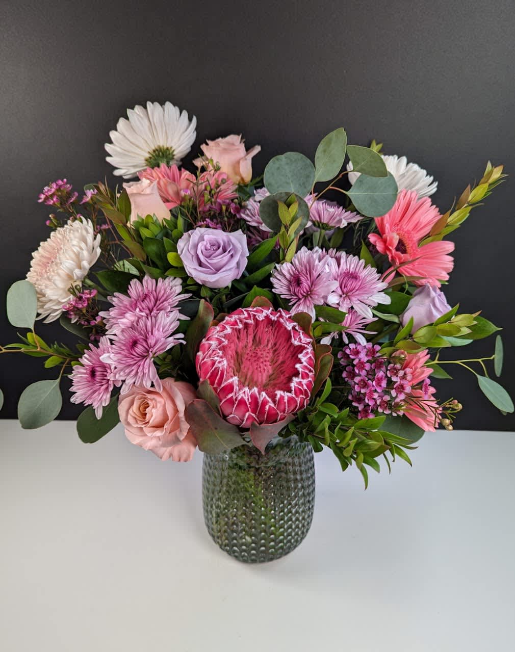 Elegant Mauve Blooms - Explore the Elegance of Mauve Blooms Floral Arrangement  Mauve Blooms showcases the soft, muted elegance of mauve flowers, weaving together a harmonious tapestry of delicate hues in a timeless floral arrangement. This arrangement features:  -Pink protea -Pastel pink and lavender spray mums -Peach mums -Hot pink gerbera daisies -Pink and lavender roses  Indulge in the enchanting beauty of this bouquet, perfect for adding a touch of sophistication to any setting.