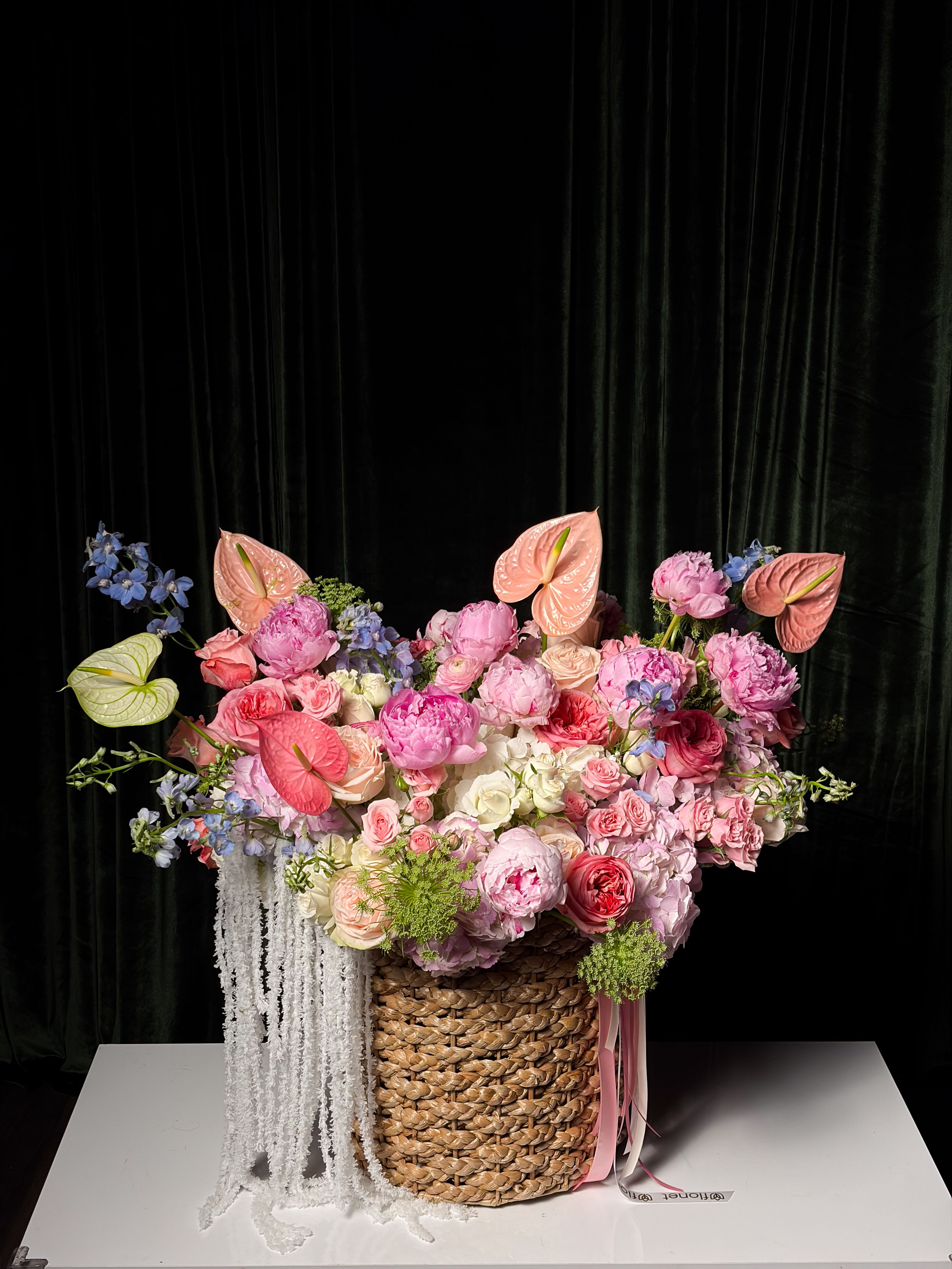 Pink Sonata Basket - A soft, romantic composition that feels like a quiet love song. Pink Sonata is built around lush, garden-style blooms in delicate shades of blush, rose, and cream. Peonies and roses create fullness and depth, while graceful anthuriums add a modern, sculptural touch. Light blue accents and airy textures soften the palette, and cascading white amaranthus gives the arrangement an elegant, couture finish. Designed in a natural woven basket, this piece feels both refined and organic—perfect for grand gestures, celebrations, or as a statement interior arrangement.
