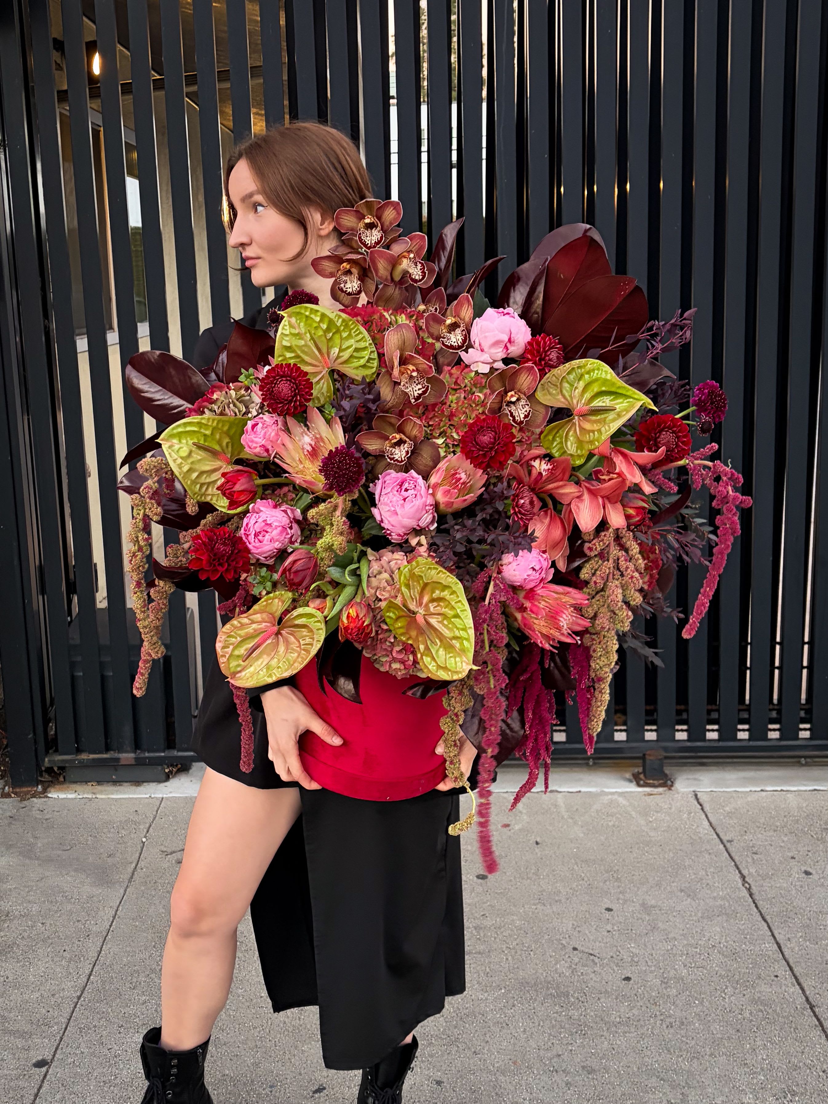 Garnet Muse - A bold, artistic arrangement with a wild, editorial spirit. Garnet Muse blends deep burgundy, wine, and antique pink tones with unexpected green accents, creating a rich, layered composition that feels both powerful and sensual. Sculptural anthuriums and orchids set the mood, while roses, textured blooms, and cascading amaranthus add depth, movement, and drama. Designed in a vibrant red hatbox, this piece feels like wearable art—confident, expressive, and unapologetically statement-making.