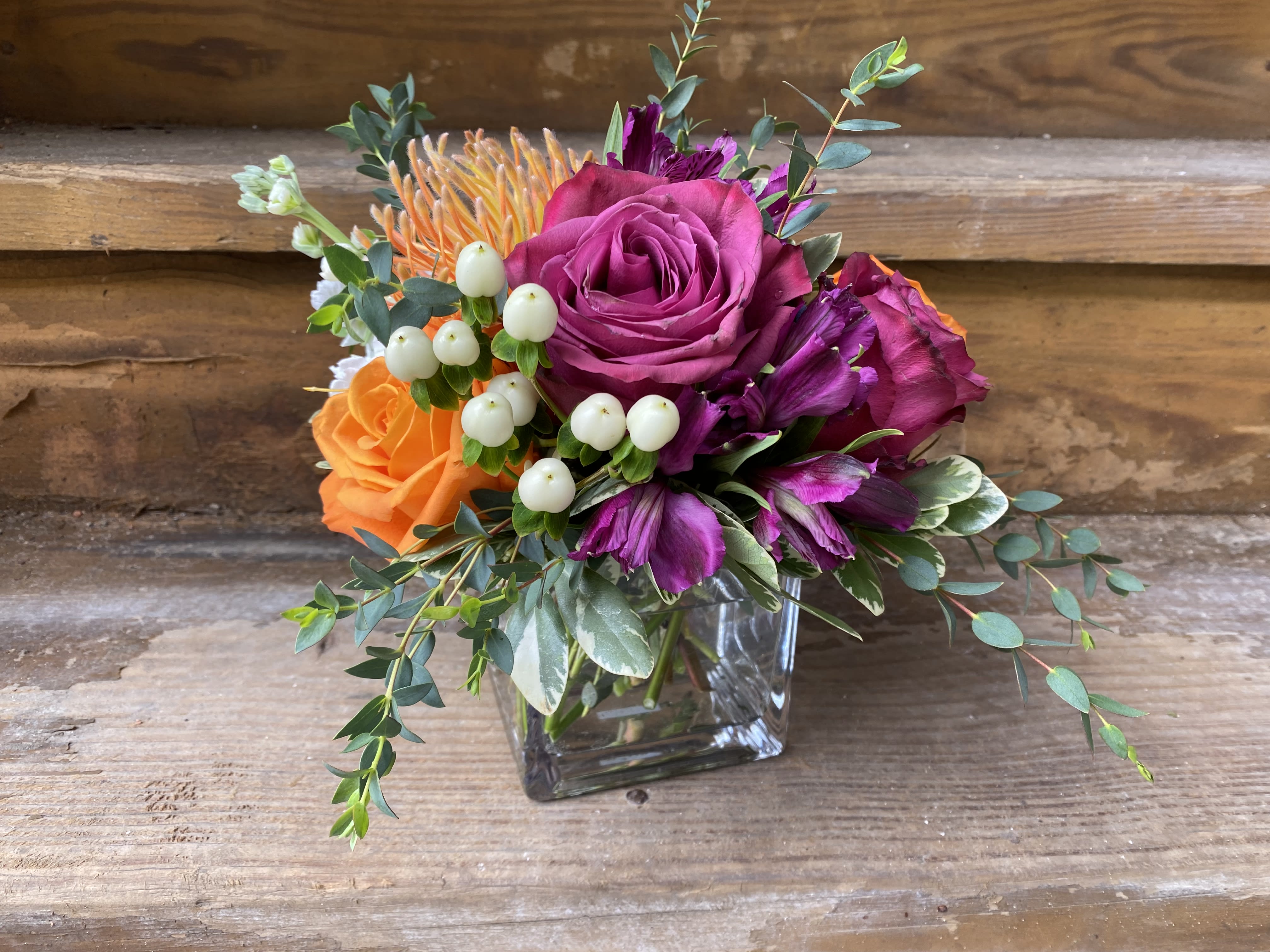 Nirvana - low arrangement - Low Arrangement in a Clear Glass Cube of jewel tone roses, pin cushion, alstromeria, stock, mums, berries and flowing greens.