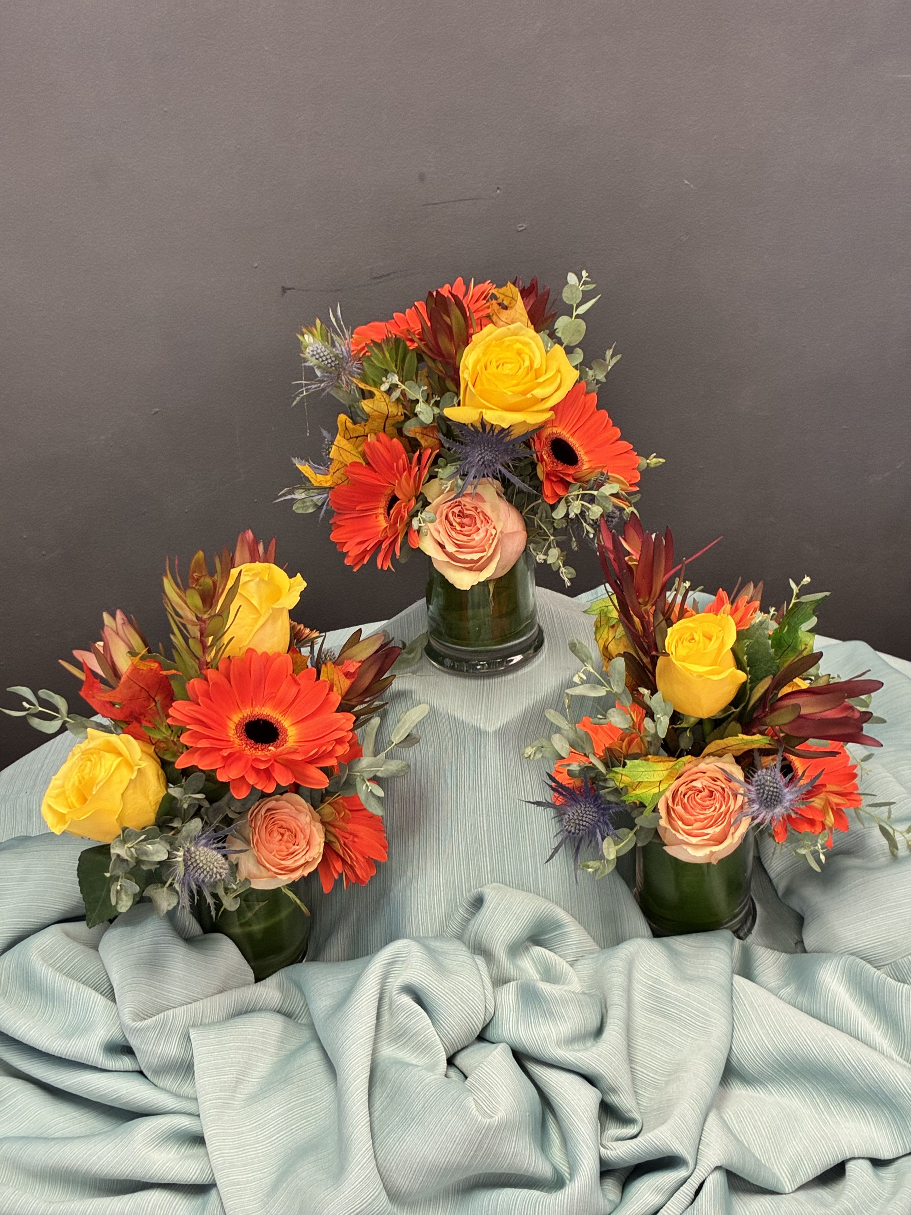 Amber Breeze  - petite arrangements glow with sunset-inspired warmth and texture. Each green glass vase holds a cheerful mix: yellow and peach roses, orange gerbera daisies, and blue thistle for a touch of contrast. Red foliage and lush greenery add depth, while the soft blue fabric beneath them evokes sky meeting earth. Together, they feel playful yet polished—perfect for intimate gatherings, seasonal tablespaces, or brightening a quiet corner with autumn charm. great for individual gifts!