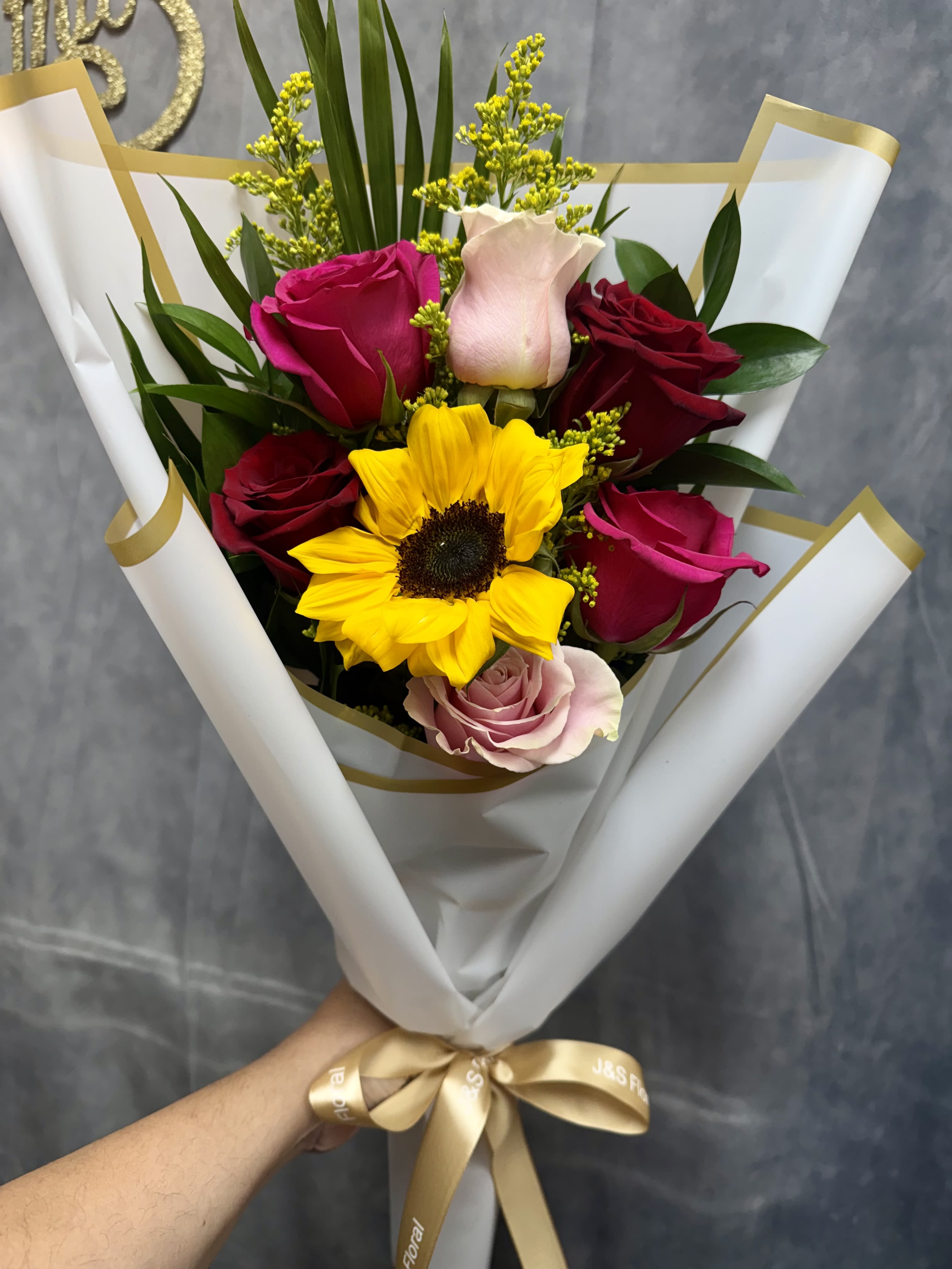 Sun Kissed Romance Bouquet  - A radiant mix of cheerful sunflower and romantic roses in shades of red, blush, and pink, accented with fresh greenery and golden filler blooms. Wrapped in elegant white and gold, this bouquet captures warmth, love, and a touch of sunshine—perfect for brightening someone’s day or celebrating a special moment.