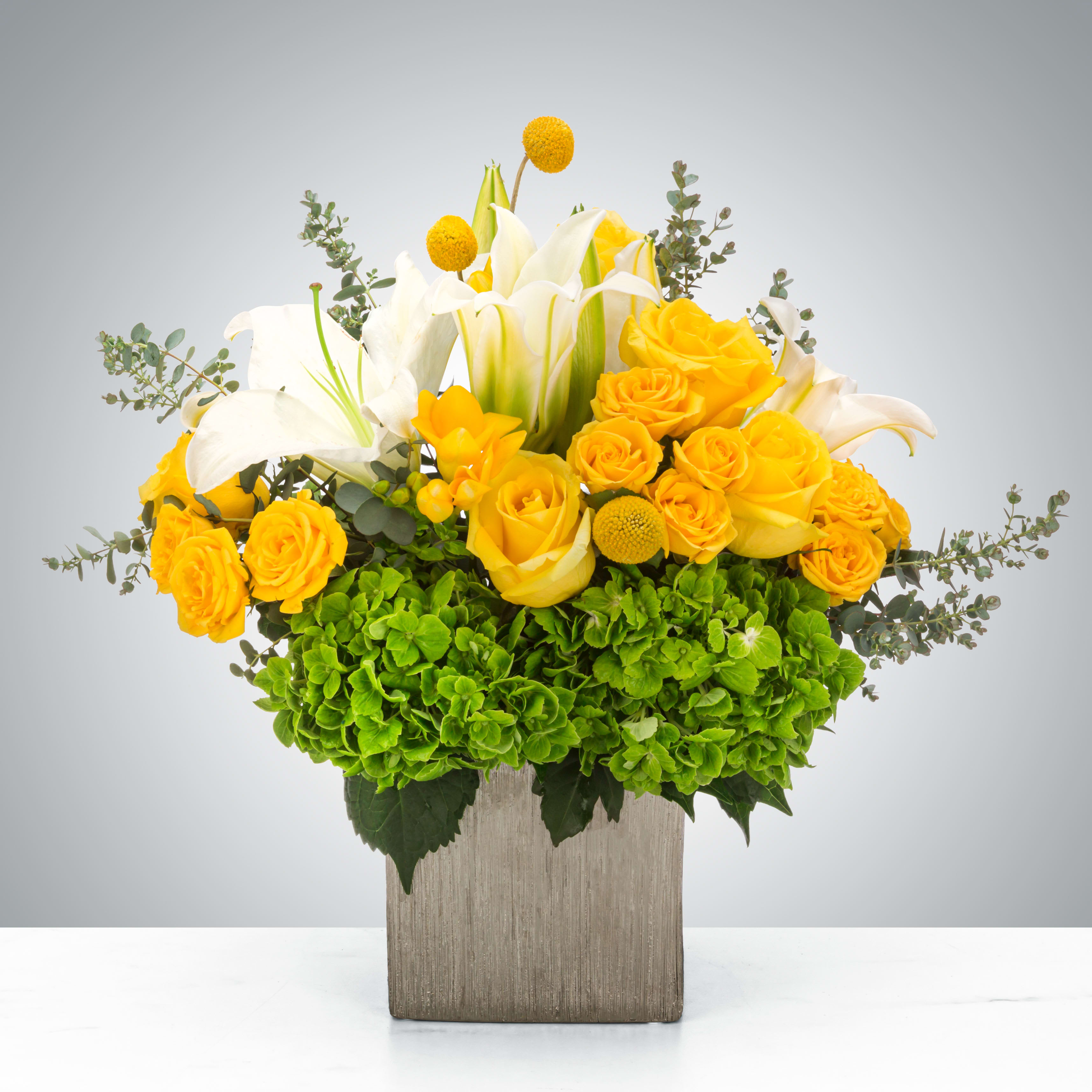 Silver Sunshine  - This arrangement features a luxury vase filled with white lilies and yellow roses with eucalyptus and hydrangea. A perfect for a 25th silver anniversary (or any anniversary really) or celebrating something special like graduation.  Approximate Dimensions: 14"D x 16"H