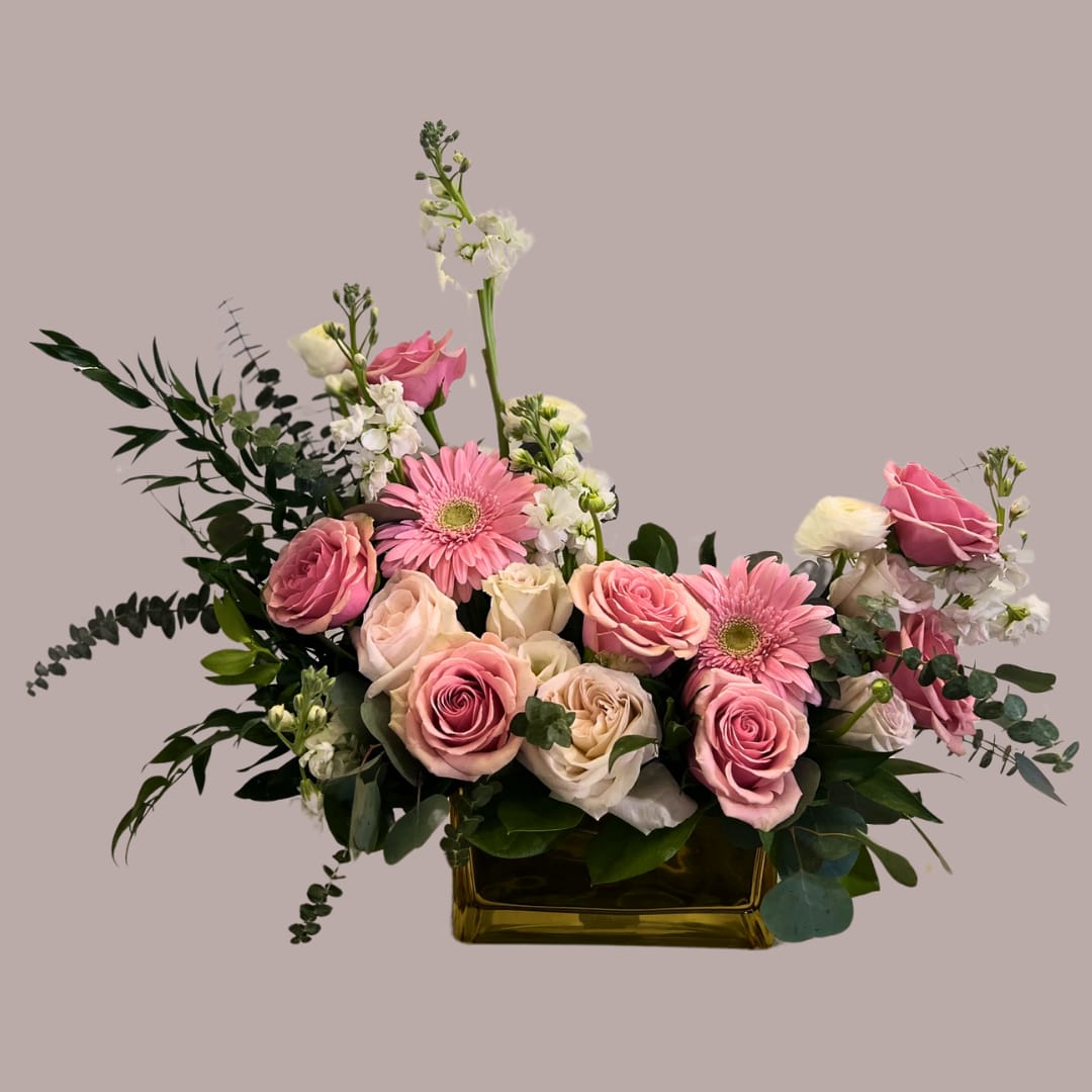 Love Song VAL2504 - Love Song VAL2504 This floral arrangement bursts with soft elegance and heartfelt charm—perfect for expressing Love on Valentine’s Day or Mother’s Day. Featuring delicate pink roses, cheerful pink gerbera daisies, and creamy white roses, the bouquet is enriched with airy white filler flowers and lush green foliage, including fern-like leaves and eucalyptus. The symmetrical composition in a sleek golden container adds a touch of sophistication, making it ideal for gifting or decorating with romantic flair. The blend of pastel blooms, textured greens, and warm metallic accents creates a graceful, garden-inspired centerpiece that speaks volumes through its floral beauty.