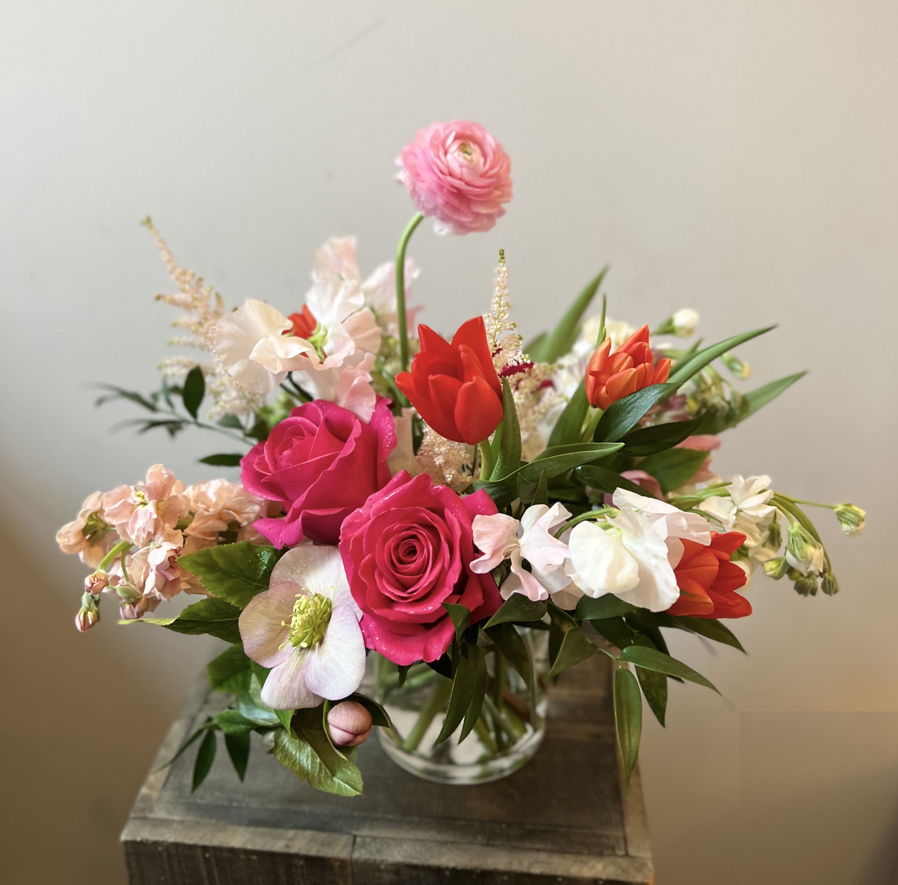 Under the Parasol - A sweet and lovely arrangement of early springtime blooms, evoking a whimsical garden stroll under a twirling parasol.  Sweet peas, tulips, ranunculus, and more fill this 4x4-inch glass cylinder vase.