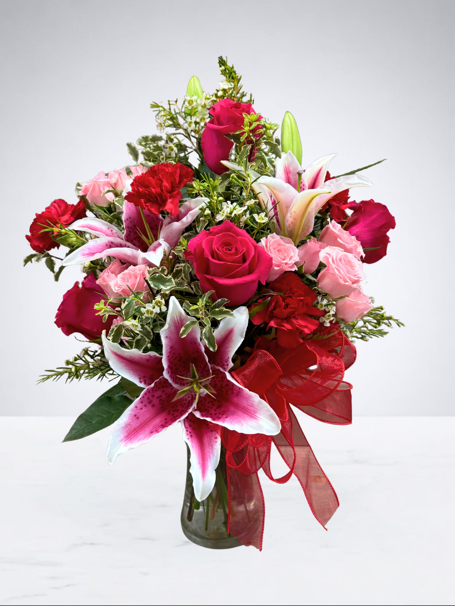 Blushing  - This stunning arrangement is a beautiful celebration of love and elegance. Featuring a lush mix of soft pinks, rich reds, and vibrant greens, it’s designed to make a heartfelt statement. Accented with graceful blooms and finished with a flowing red ribbon, this arrangement is perfect for anniversaries, Valentine’s Day, birthdays, or simply letting someone know how special they are. 