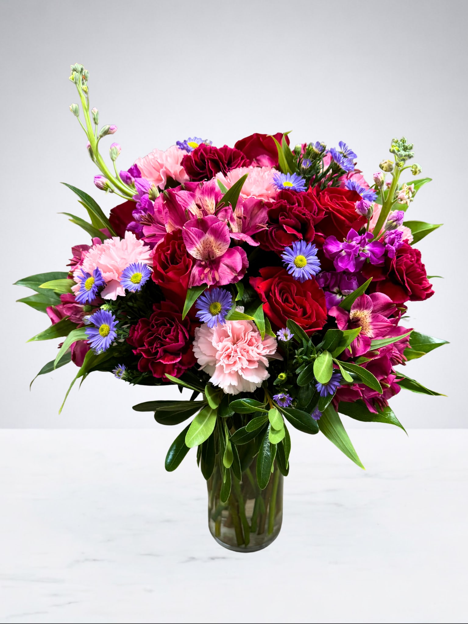 Vibrant Duet - A stunning mix of deep red roses and pink carnations accented by playful purple daisies and exotic alstroemeria. This lush, rounded bouquet is the perfect way to send a pop of color and a big smile to someone’s doorstep.