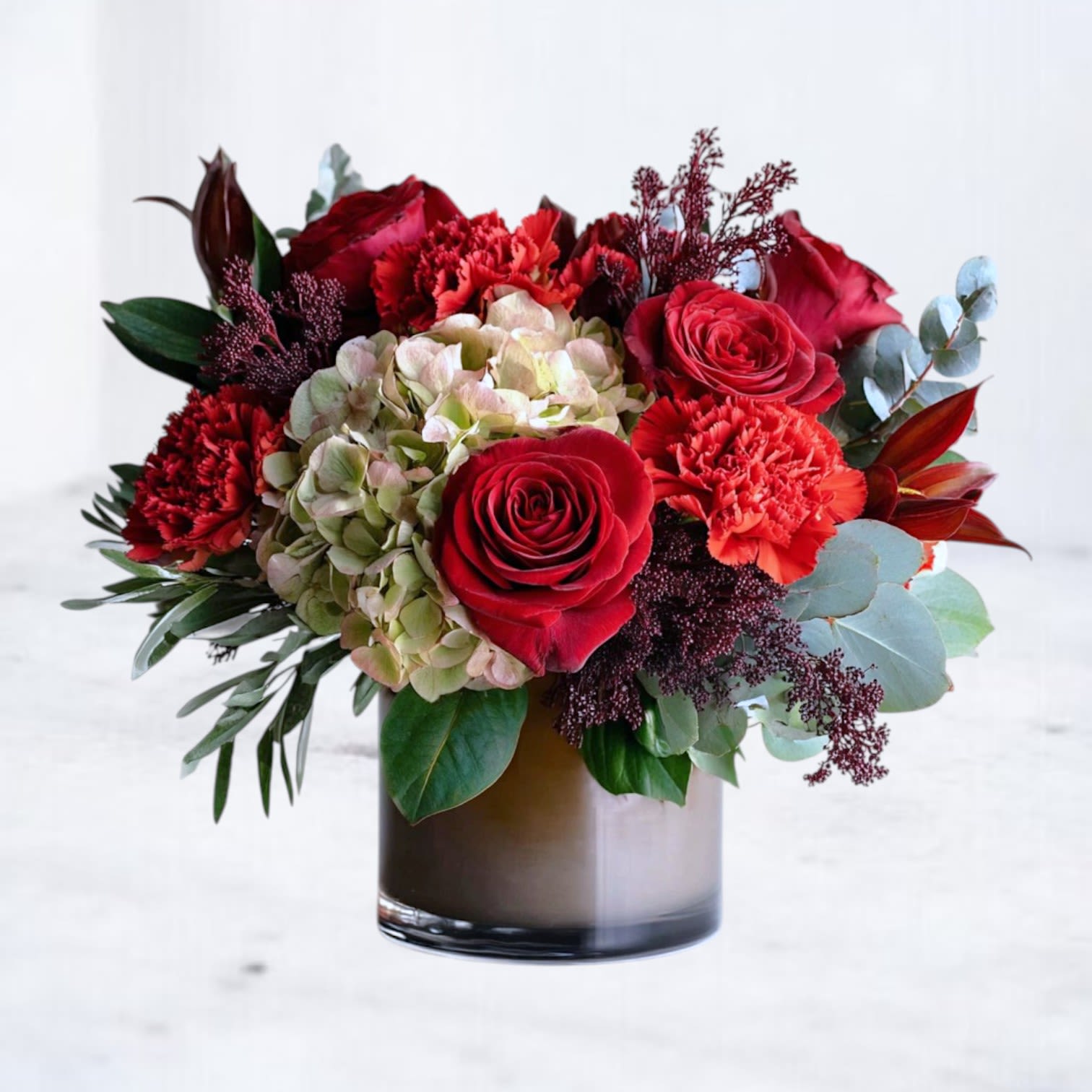 Burnished Ruby - The Burnished Ruby bouquet is a dramatic showcase of passion and elegance, featuring velvety red roses, deep crimson carnations, burgundy accents, and antique hydrangea. Arranged in a sleek glass vase, this design blends bold romance with modern sophistication — making it an unforgettable gift for anniversaries, birthdays, or everyday luxury gestures.  Available for same-day flower delivery in Magnolia, The Woodlands, Spring, and Montgomery, this bouquet captures the warmth of love and the richness of jewel-tone beauty.
