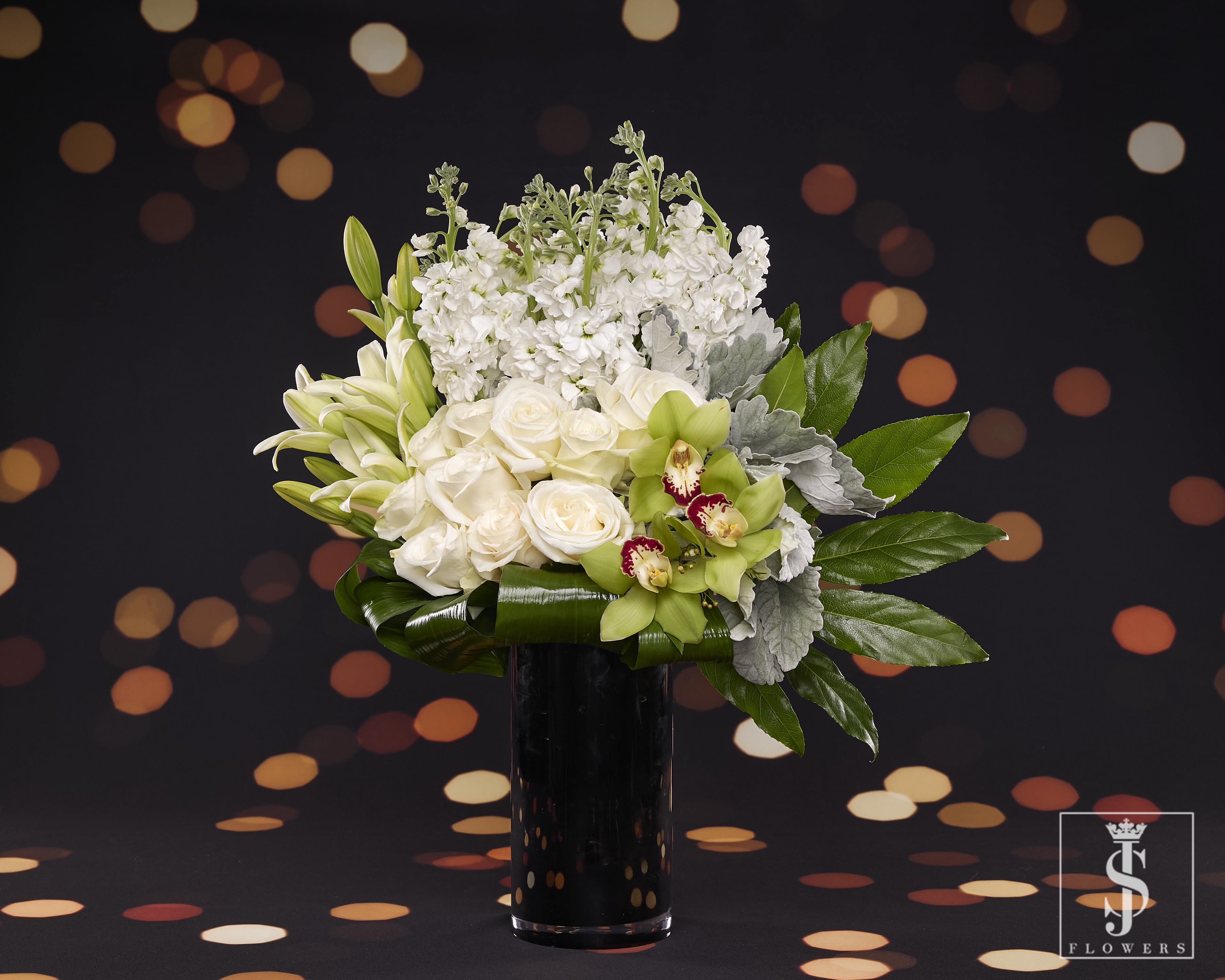 New Year Delight  - Celebrate the New Year with timeless elegance through this luxurious arrangement. Featuring pristine **white roses** symbolizing purity and new beginnings, alongside elegant **white lilies** that represent renewal and celebration, this design radiates sophistication. Accents of exotic **green cymbidium orchids** add a refreshing pop of color, while delicate **stock flowers** bring a lush and full texture, symbolizing happiness and contentment. The arrangement is completed with frosty **dusty miller foliage** and vibrant **glossy green leaves**, creating a harmonious blend of freshness and grace. Perfect for adding a touch of refined beauty to your New Year's festivities!