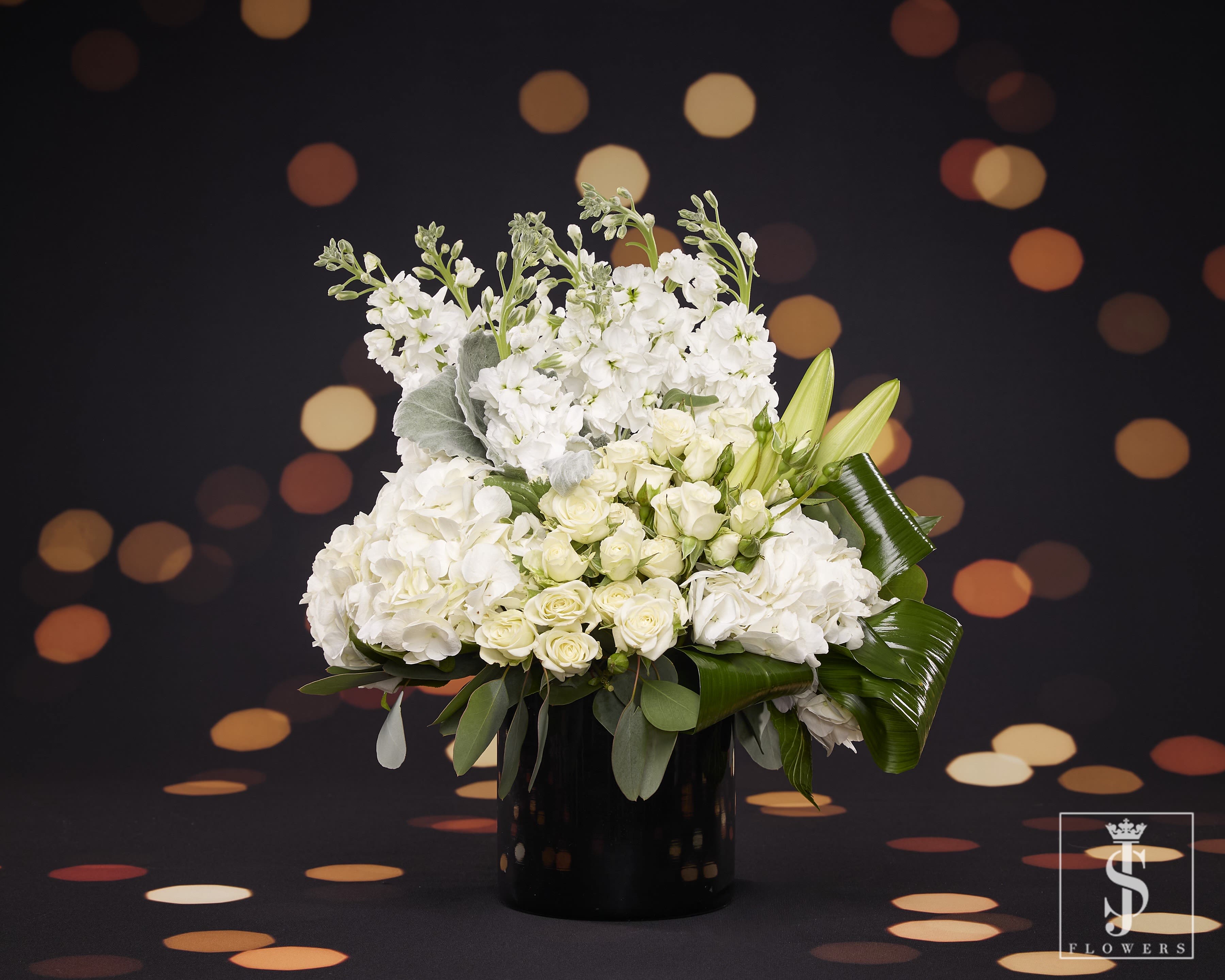 French Kiss  - Welcome the New Year in style with this captivating floral masterpiece. A grand display of white roses and spray roses, representing purity and fresh beginnings, combined with white lilies and stock flowers, signifying renewal and celebration. Soft dusty miller foliage and white hydrangeas add a frosty elegance and fullness to the arrangement, while lush green accents provide a striking contrast. Arranged in a sleek black vase, this design embodies sophistication and grace, making it the perfect centerpiece for New Year’s Eve or any celebration where elegance takes center stage.