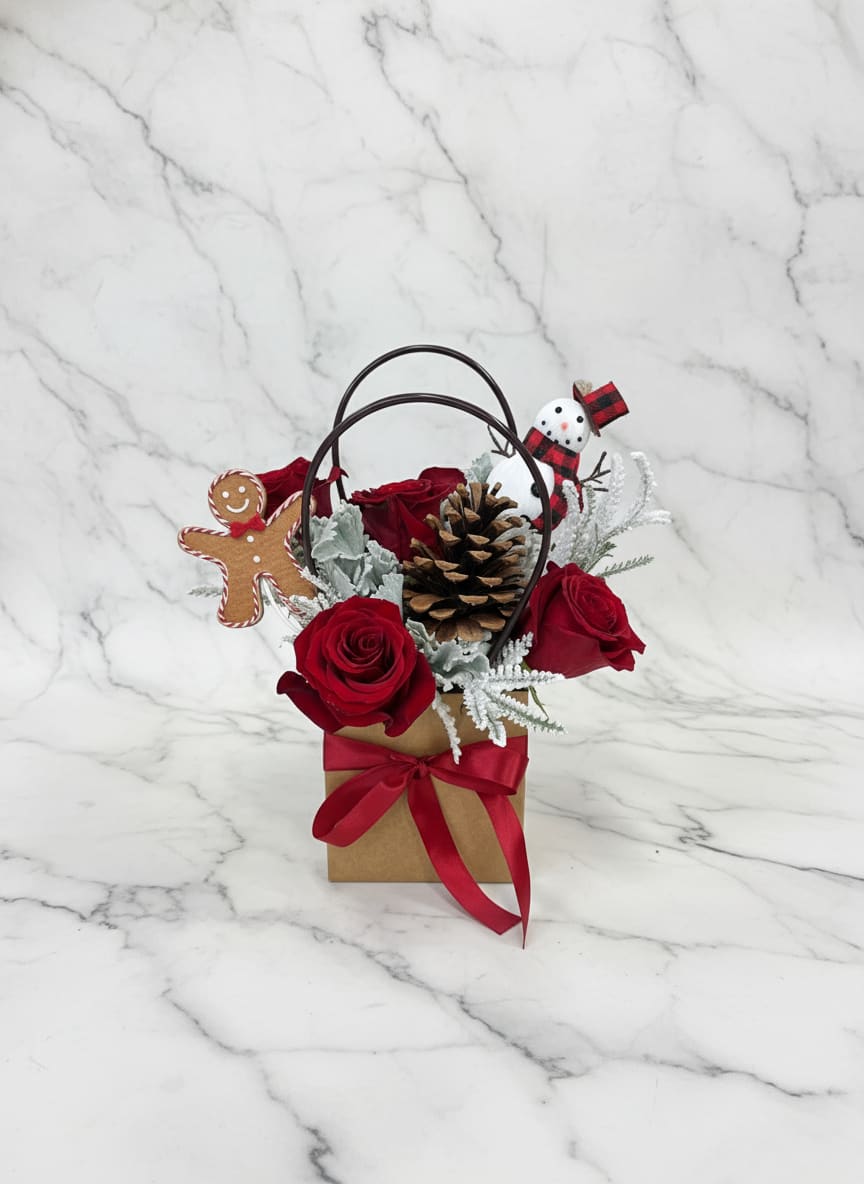 Frosted gingerbread bag  - A charming winter arrangement designed in a rustic square kraft paper bag, featuring classic red roses nestled among flocked winter greenery and soft dusty miller. Finished with natural pinecones, a cheerful snowman, and a sweet gingerbread man, this design brings a cozy, festive touch of winter whimsy to any space.