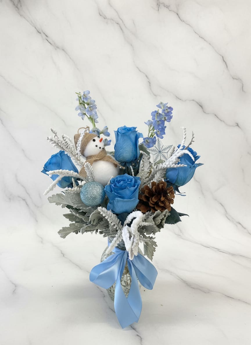 Frosty the snowman vase - A frosty the snowman vase features soft baby blue roses paired with silvery dusty miller and flat winter greenery. Accented with a natural pinecone and a cheerful snowman, this arrangement captures the crisp beauty of winter with a playful, seasonal touch.