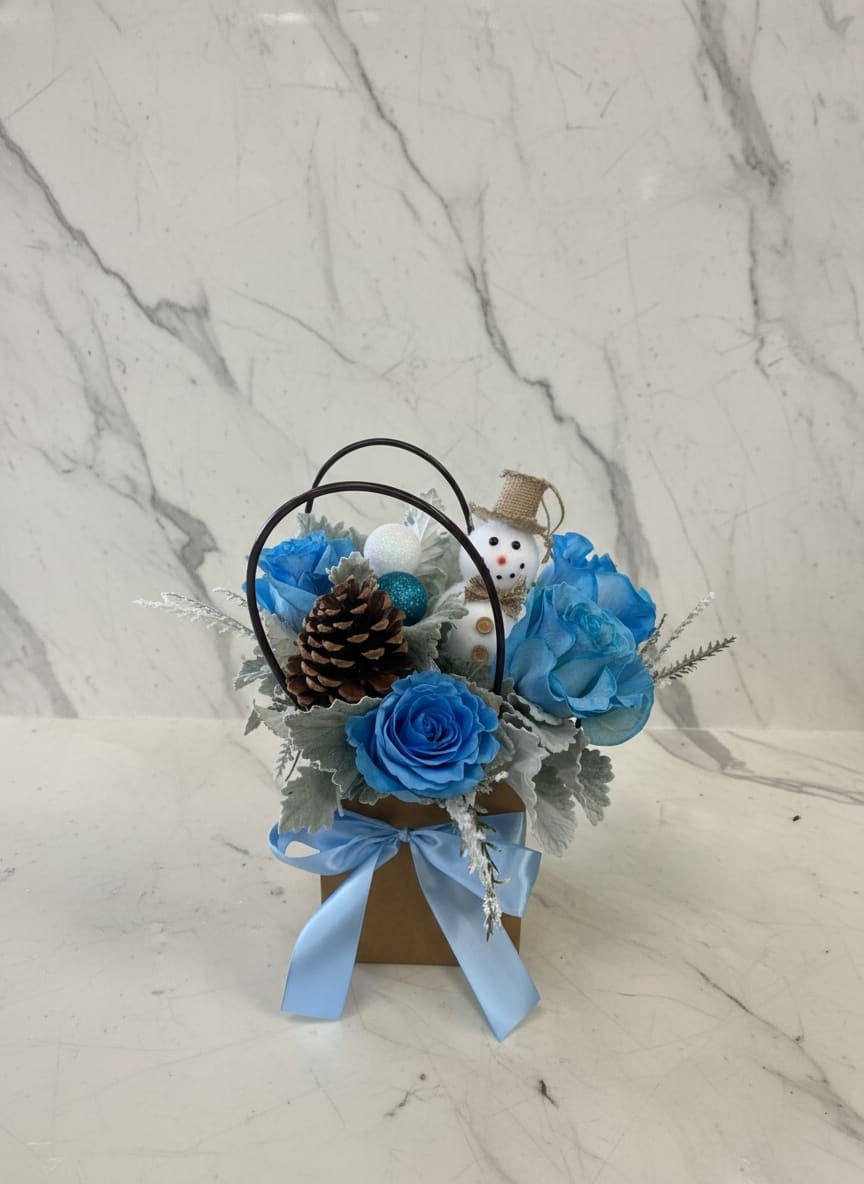Frosted winter bag  - A frosty winter arrangement designed in craft Bag, featuring soft baby blue roses surrounded by silvery dusty miller and flat winter greenery. Finished with a natural pinecone and a cheerful snowman accent, this design captures the crisp charm of a snowy winter day with a playful seasonal touch.