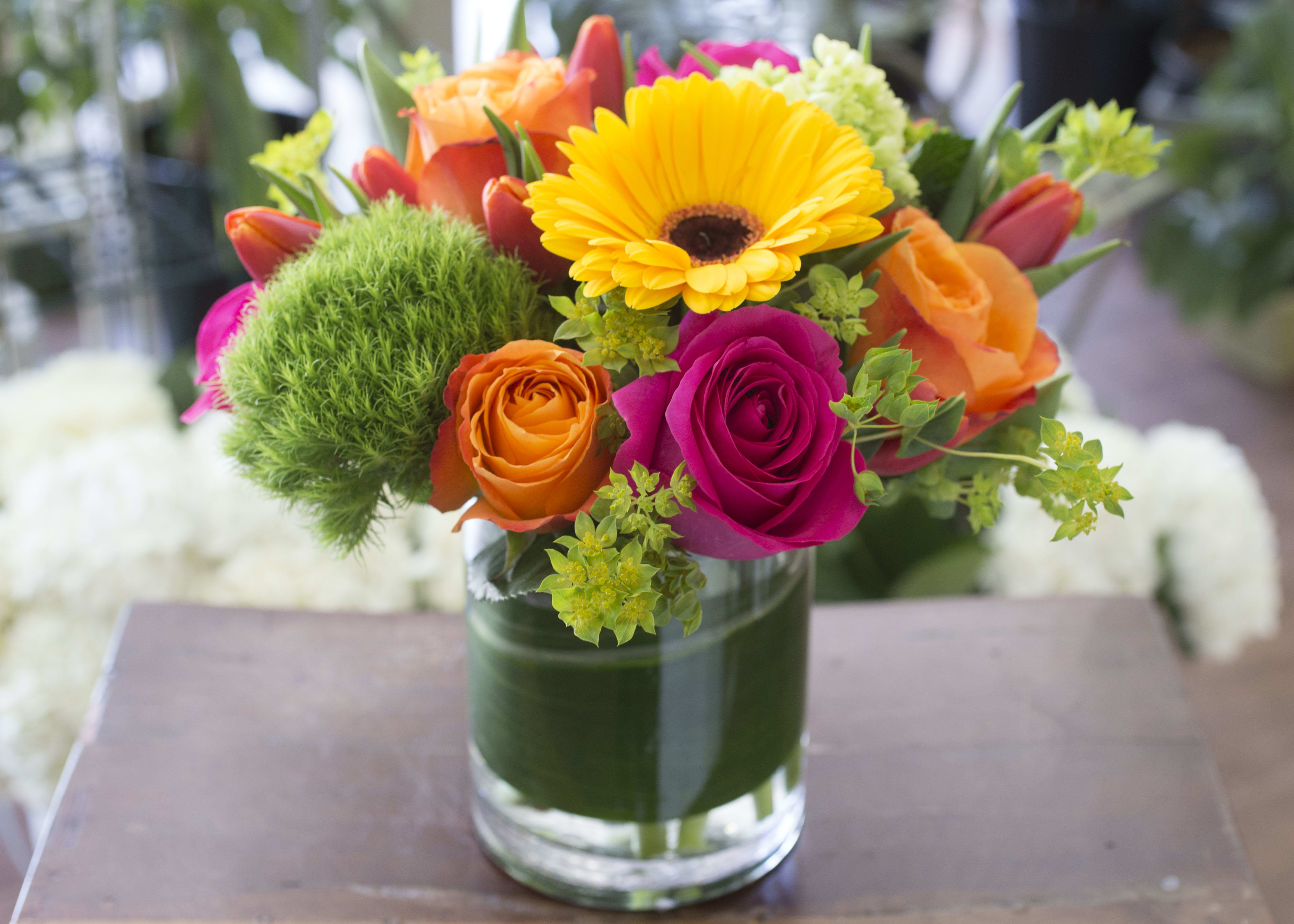 Caroline - Bright colors of Gerbera Daisies and Roses with seasonal accents arranged in a clear glass vase.