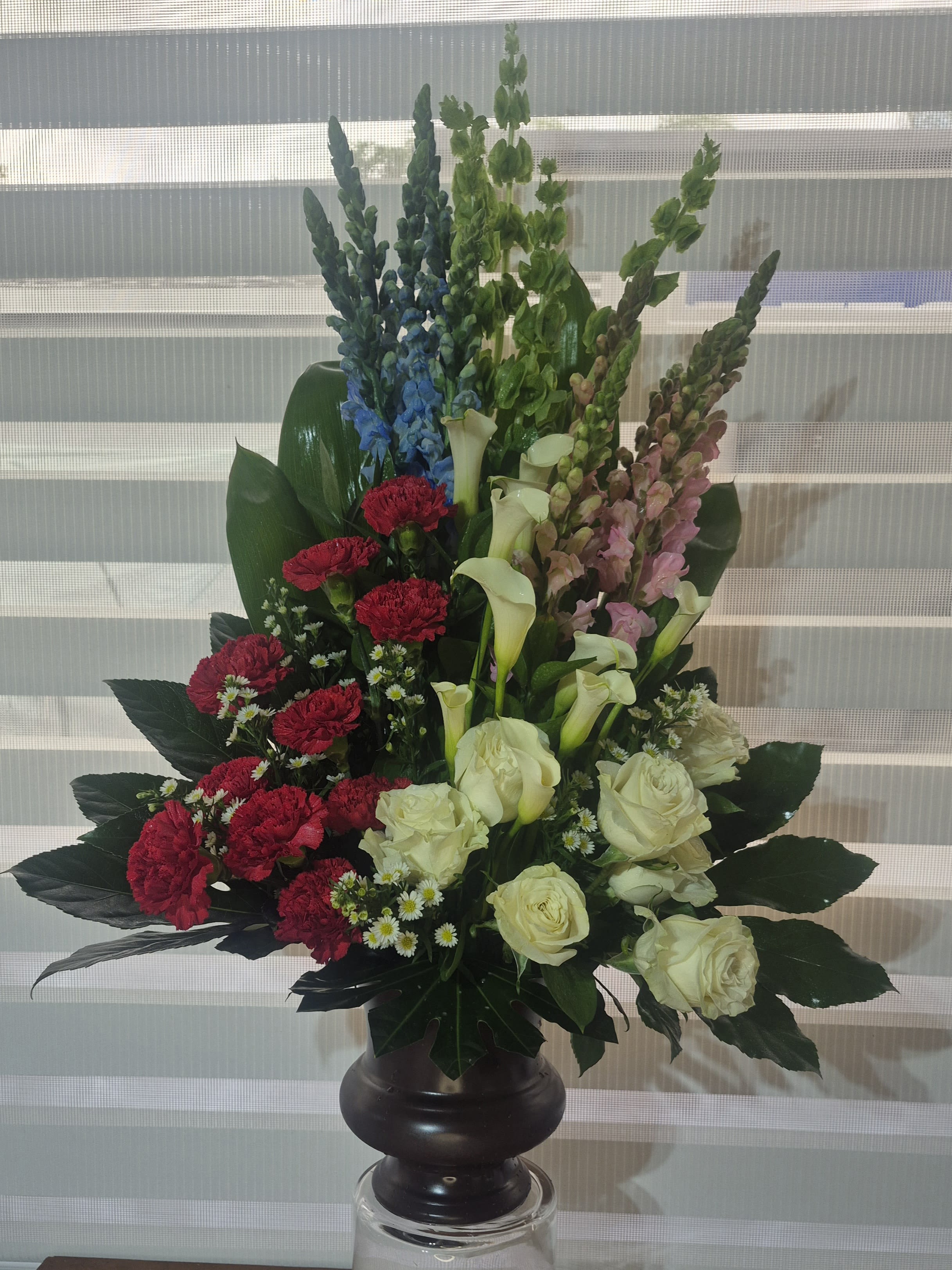 PEACEFUL REST - A ONE SIDED ARRANGEMENT CONSISTING OF WHITE ROSES, WHITE MINI CALLA LILLIES, PINL SNAPDRAGONS, RED CARNATIONS, WHITE MONTI-CASINO FILLER, BLUE DELPHINIUMS AND BELLS OF IRELAND FLOWERS, ARRANGED IN A URN SHAPED VASE.