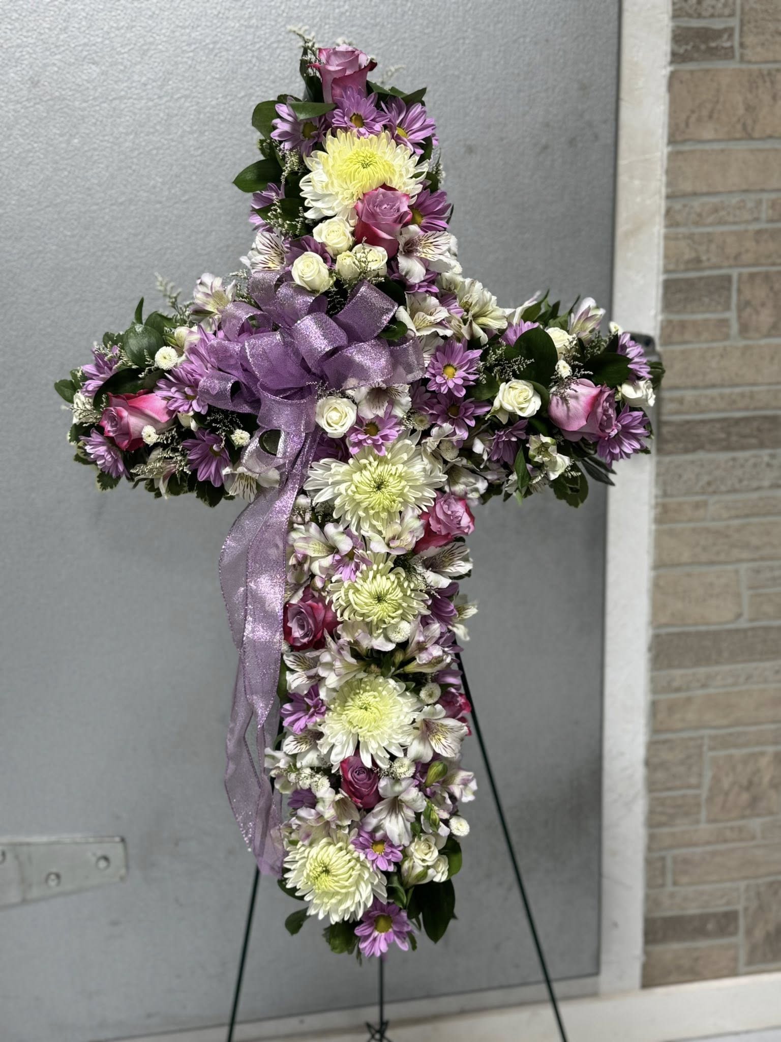 Lavender Grace Cross Standing Spray  - The Lavender Grace Cross Standing Spray is a beautiful expression of faith, peace, and remembrance. Designed in the shape of a cross, this standing spray features soft lavender blooms complemented by fresh white florals and lush greenery, symbolizing grace, devotion, and eternal rest. The soothing lavender tones convey calm and reflection, while the white flowers represent purity and peace.  This floral cross spray is a meaningful tribute for funeral services, memorials, church services, or graveside ceremonies. Handcrafted with care by our professional florists, the Lavender Grace Cross Standing Spray offers comfort to grieving families and serves as a lasting symbol of love and faith.  Details:  Cross-shaped standing funeral spray  Lavender and white floral design with greenery  Appropriate for funerals, memorial services, and religious services  Delivered fresh and arranged by a local florist