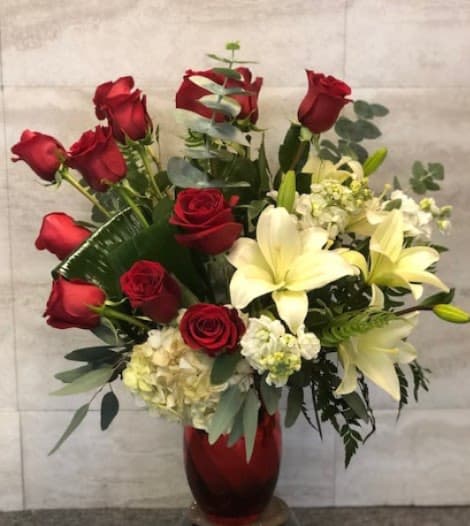 love... forever - A classic floral arrangement displayed in a red glass vase. It features deep red roses paired with large, creamy white lilies, soft white hydrangea, and smaller white accent flowers. Lush green foliage fills out the bouquet, giving it a full, elegant look. The contrast between the rich red roses and the light-colored flowers creates a romantic and refined appearance, suitable for a formal setting or special occasion. 