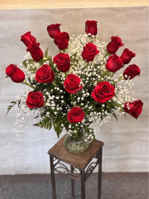 18 red roses - A tall, elegant bouquet of red roses arranged in a clear glass vase. The deep red roses are evenly spaced and surrounded by delicate white baby’s breath and green fern leaves, creating a balanced, airy, and timeless floral arrangement. 