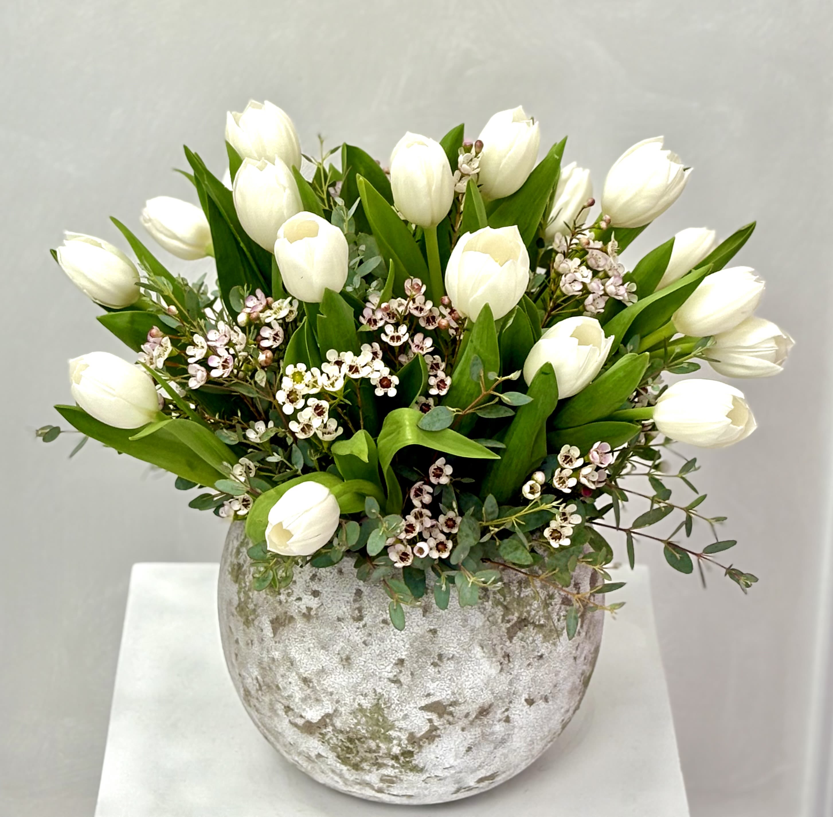 Ivory Tulip Whisper By VLM Flowers - A fresh and timeless arrangement of creamy white tulips nestled among lush green foliage and delicate wax flowers, designed in an elegant textured vase. Soft, natural, and effortlessly refined, this piece brings a sense of calm beauty—perfect for home décor, thank-you gifts, or understated celebrations.