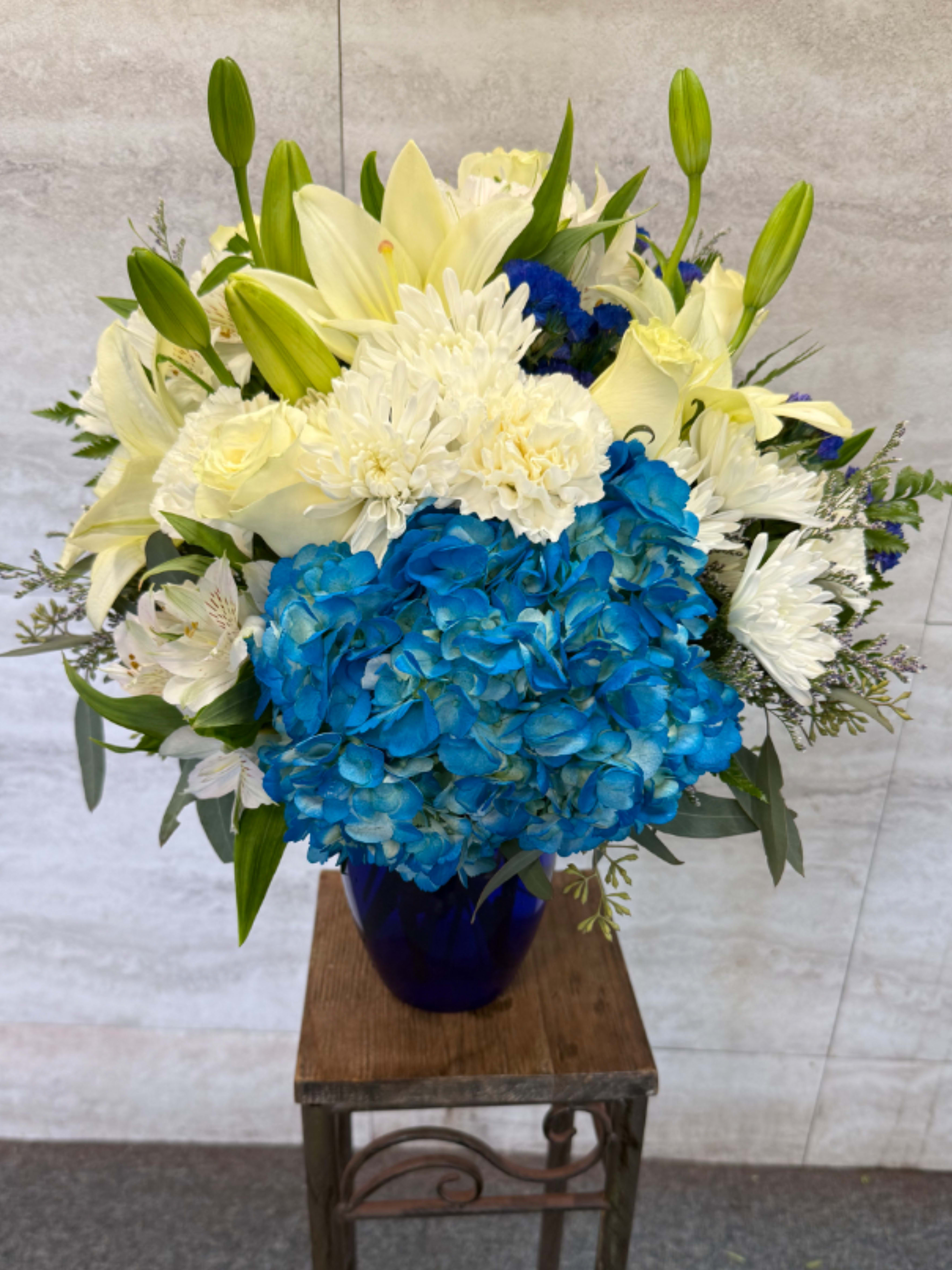 Beautiful in blue  - A floral arrangement in a deep blue vase featuring bright blue hydrangeas surrounded by white lilies, chrysanthemums, roses, alstroemeria, and statice, accented with fresh green foliage. 