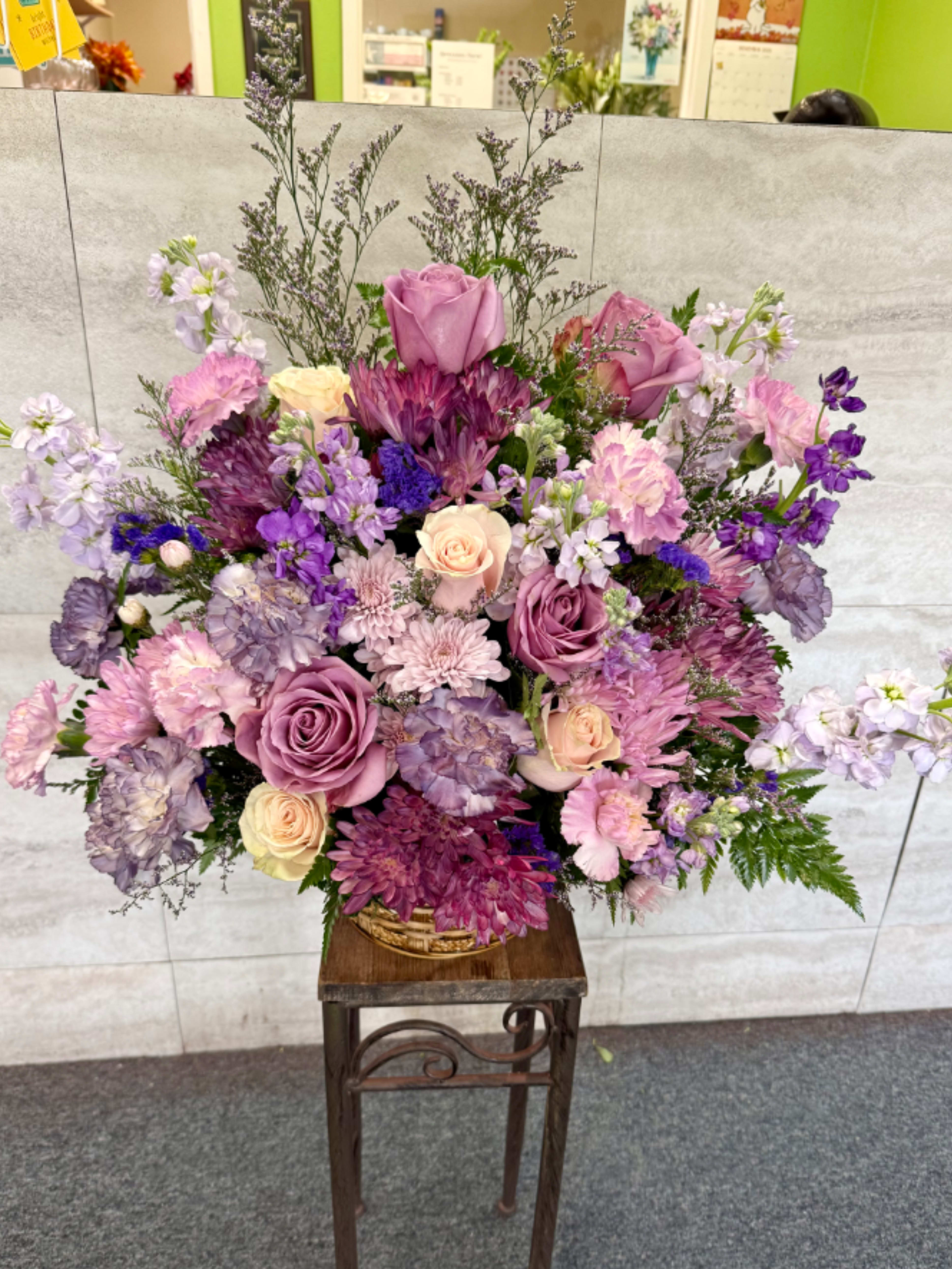 Gracious lavender's basket - This is a lush, rounded floral arrangement with a soft, romantic color palette centered on purples, pinks, and lavender tones. It features multiple varieties of roses in mauve, blush, and pale peach, paired with full chrysanthemums in dusty pink and deep plum. Ruffled carnations in lavender and light pink add texture, while delicate filler flowers in soft purple and white create an airy, layered look throughout the design.  Tall, wispy stems rise gently above the main mass, giving the arrangement height and movement, while fern greenery and fine-textured foliage frame the base and edges. The overall style feels balanced and abundant, with a mix of large focal blooms and smaller accent flowers that blend smoothly for a cohesive, elegant appearance. 