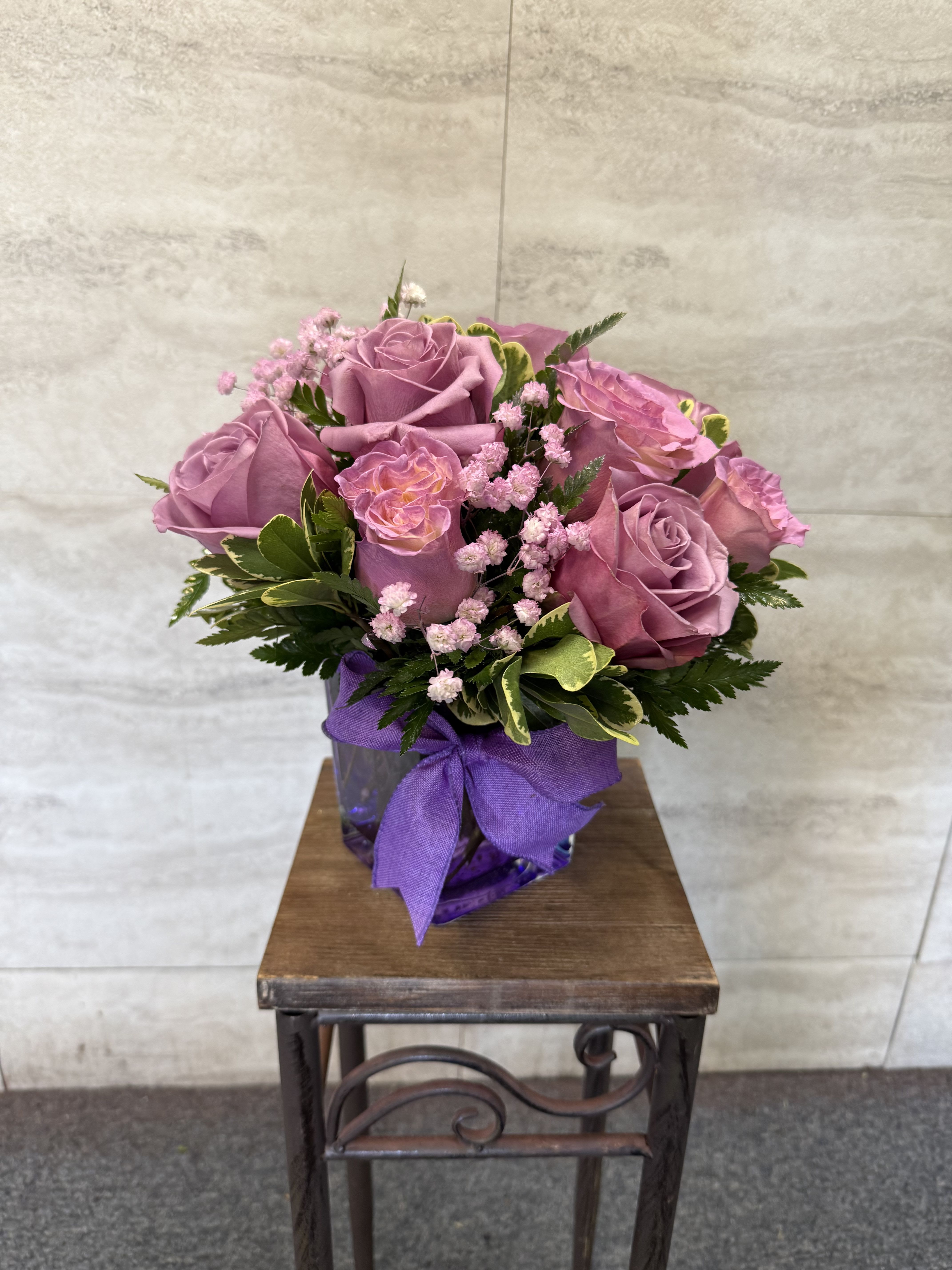 Purple haze - A bouquet of soft lavender roses with delicate pink filler flowers and greenery, arranged in a purple glass vase tied with a matching ribbon.