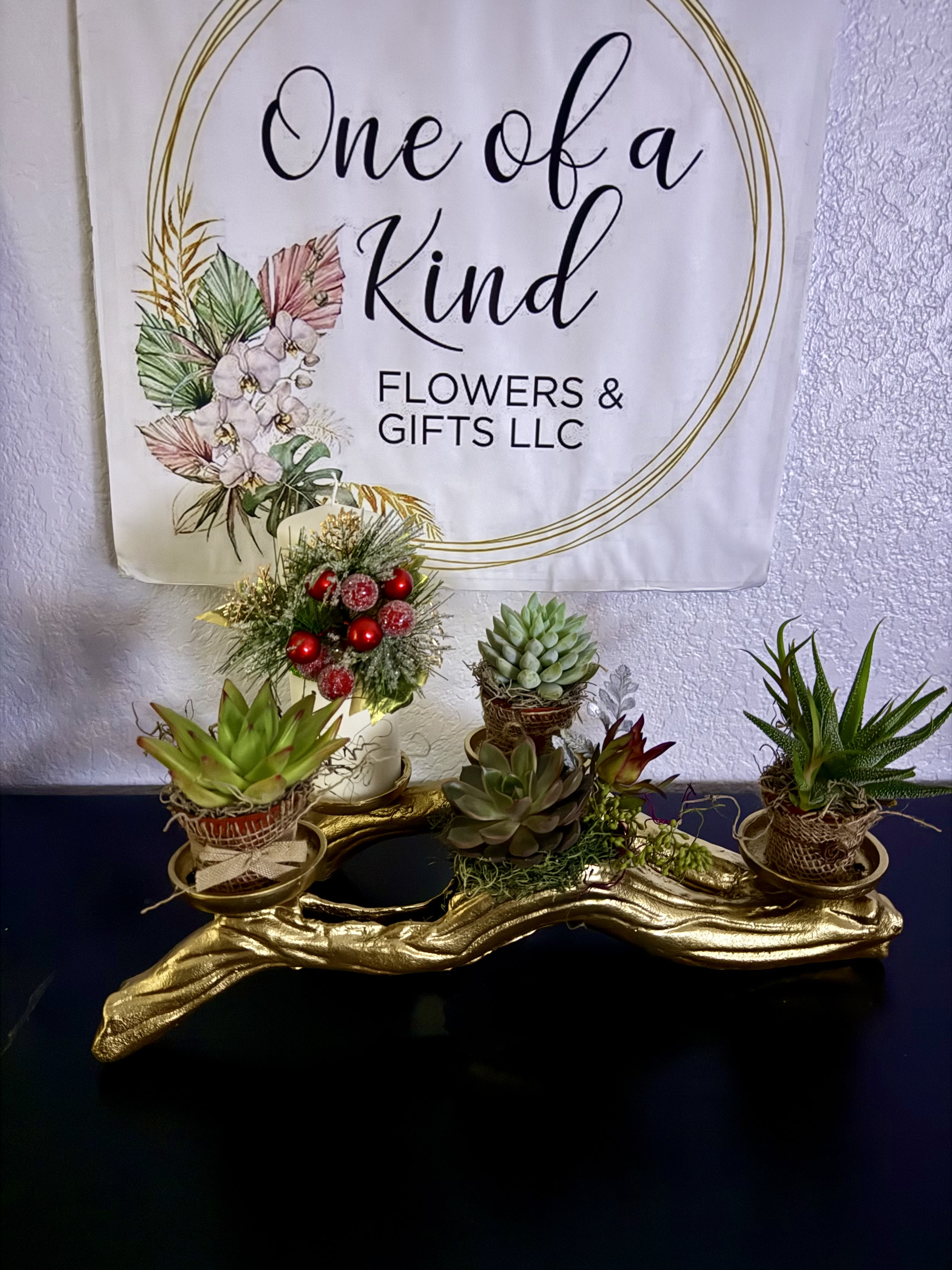 Golden Serenity Succulent Display - A luxurious gold centerpiece featuring a graceful candle surrounded by a curated selection of live succulents. Accented with festive greenery and subtle holiday details, this timeless arrangement brings warmth, elegance, and natural beauty to any space. Perfect as a sophisticated gift or seasonal décor.